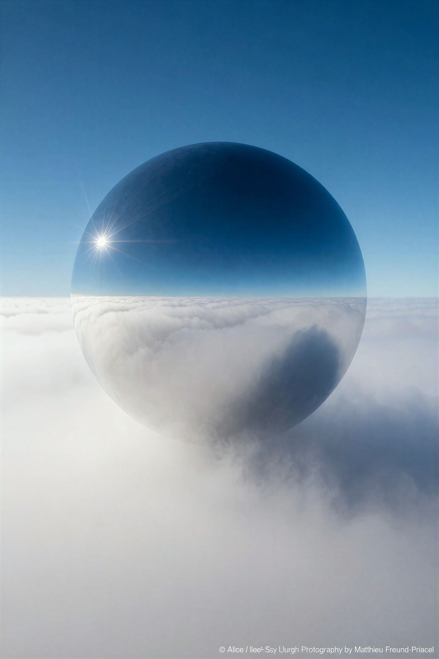 A large mirrored sphere reflects sunlight above a sea of clouds against a vivid blue sky.