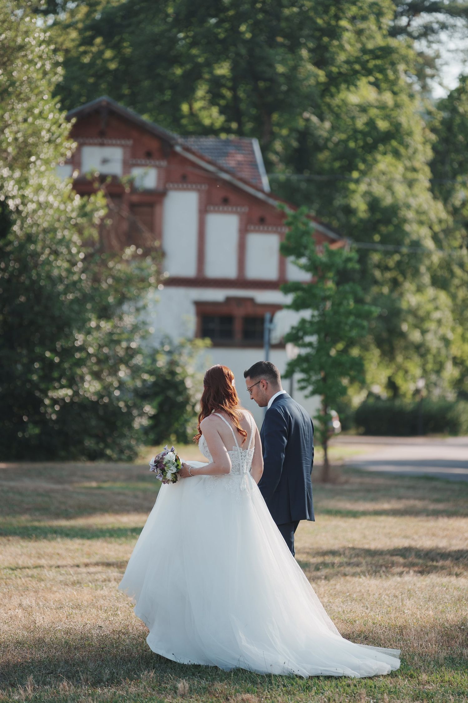 Bride in flowing white gown and groom in navy suit walk together in a lush green park.