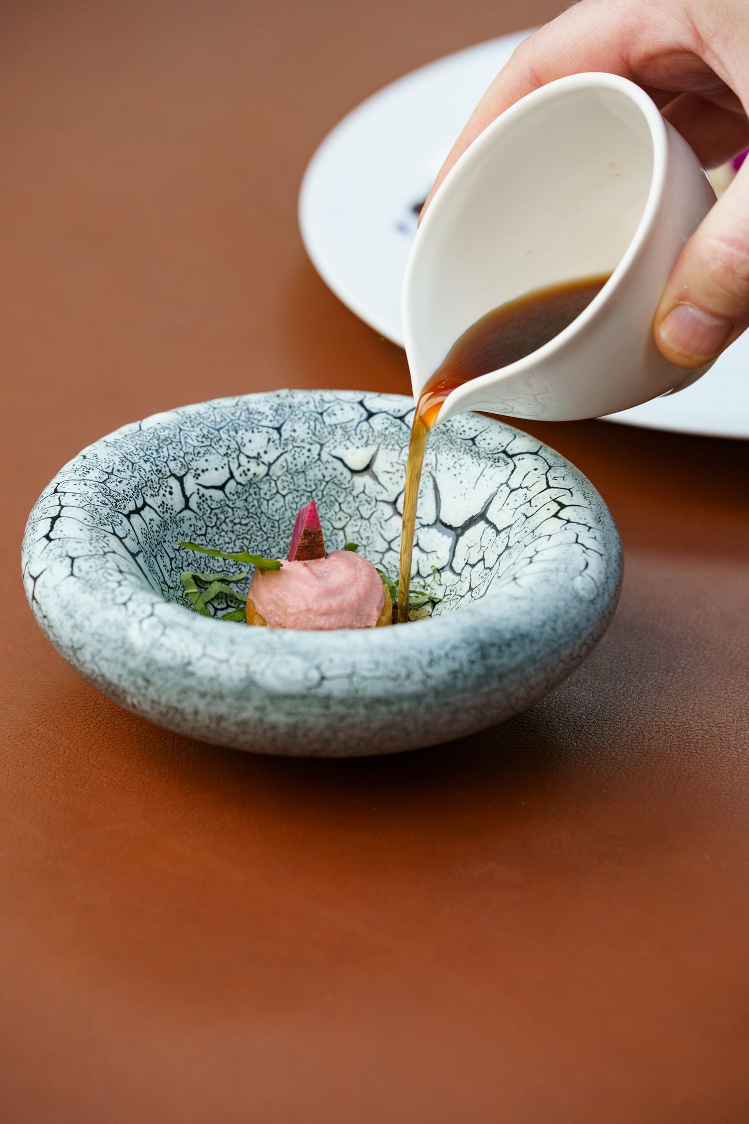 A hand pours rich brown broth into a crackle-glazed ceramic bowl with pink meat and herb garnish.