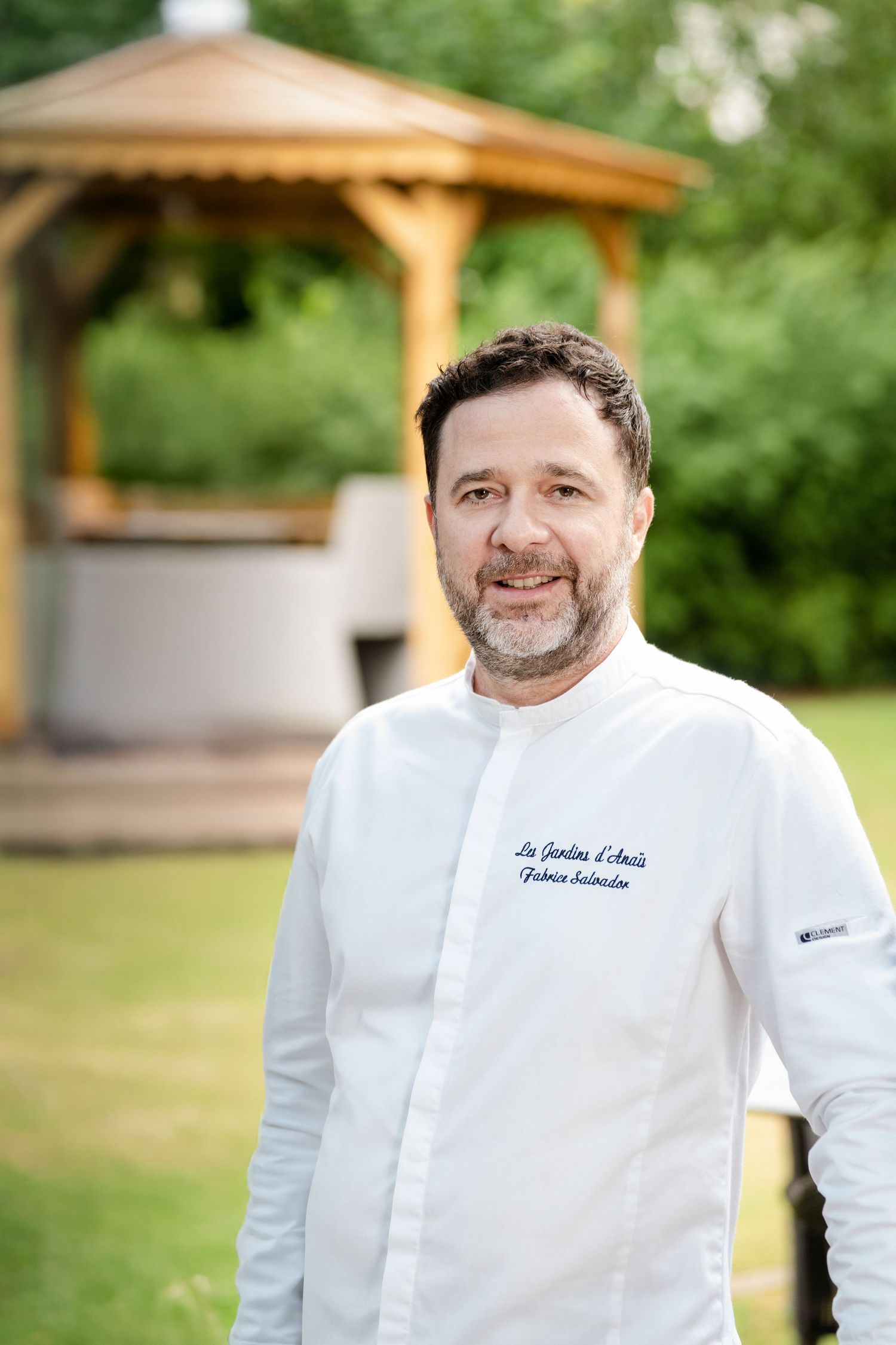 Smiling male chef in white uniform standing outdoors near a wooden gazebo surrounded by greenery.