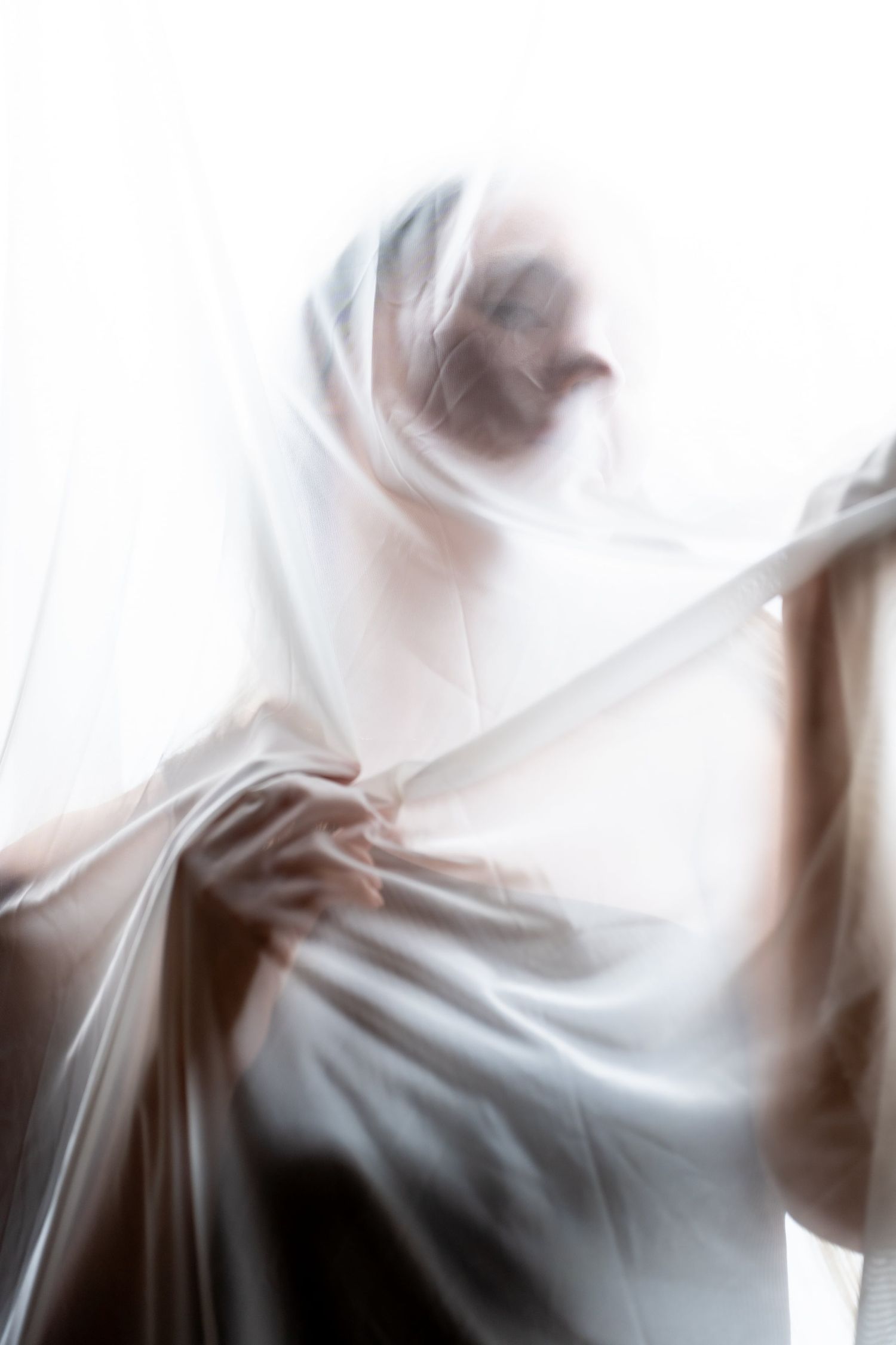 Artistic blurred portrait of a person wrapped in sheer white fabric against bright white background.