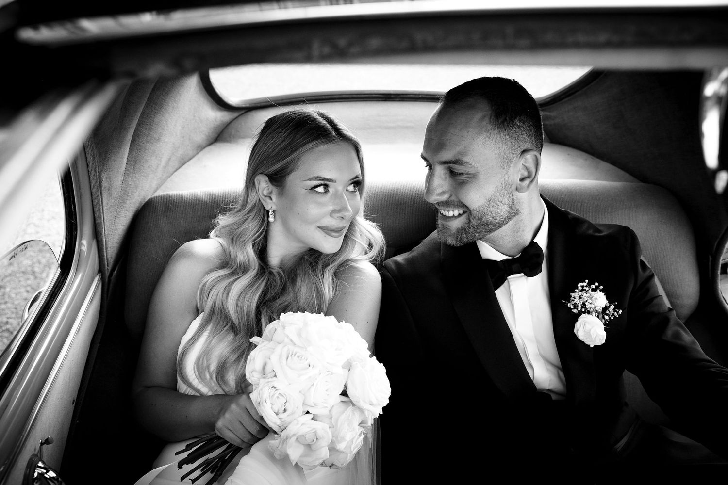 Black and white photo of smiling bride and groom sharing an intimate moment inside a vintage car.