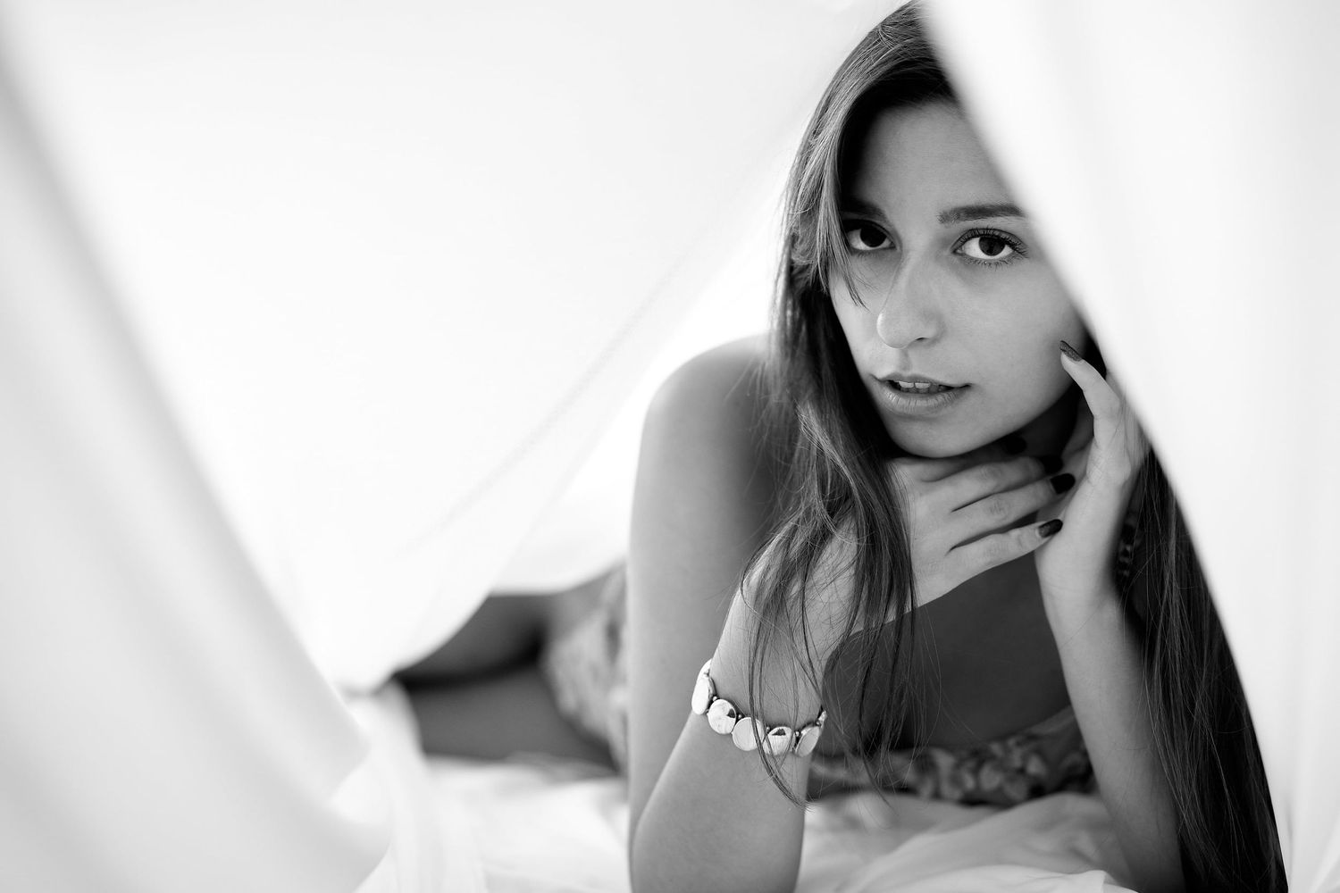 Black and white portrait of young brunette woman lying down, gazing through white fabric with bracelet.