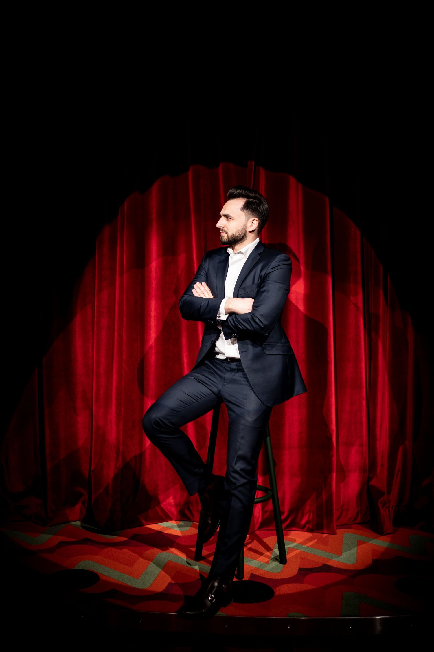 Confident man in navy suit with arms crossed standing on stage with red curtain backdrop.