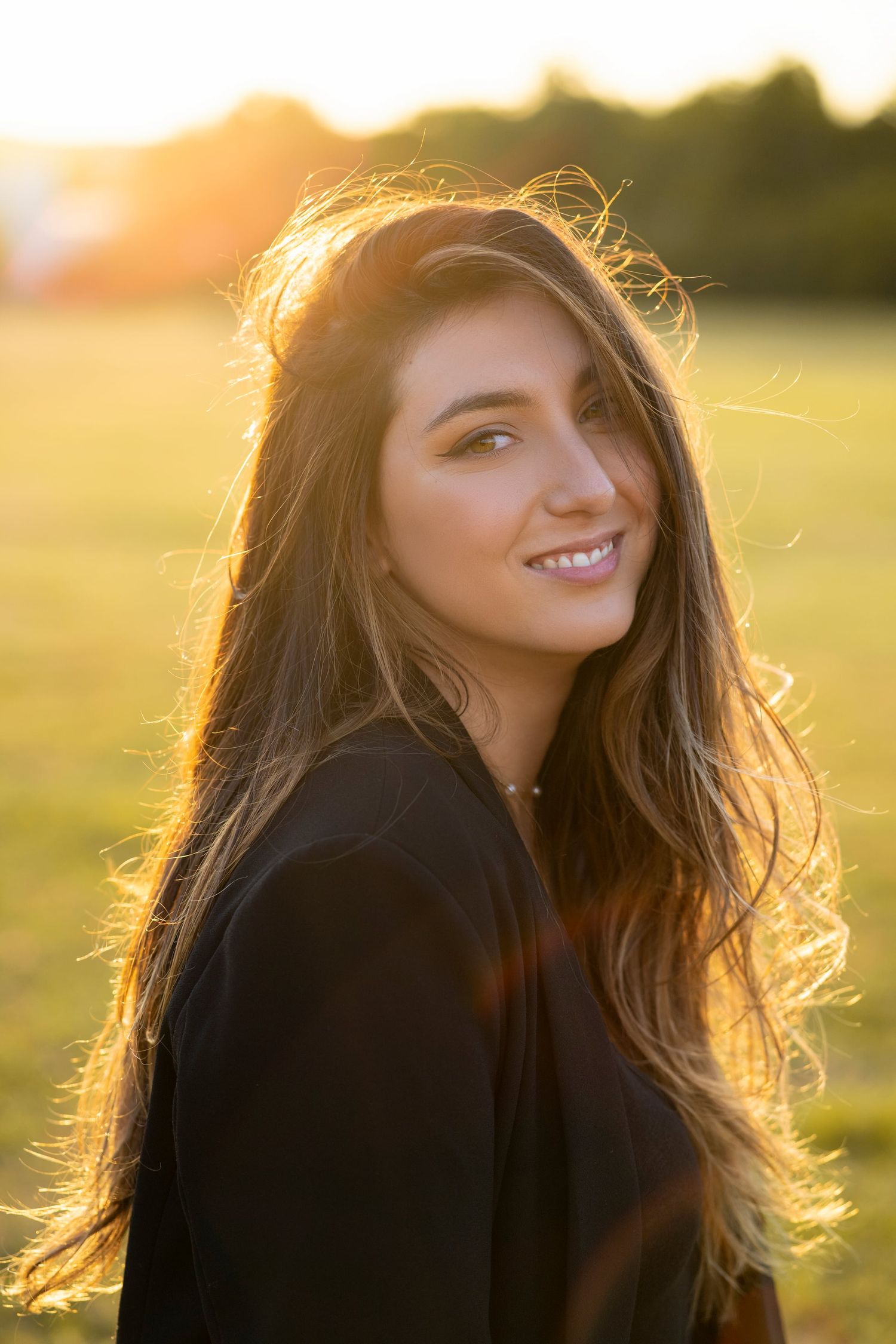 Young woman with long brown hair smiling outdoors during golden hour sunset in a black top.