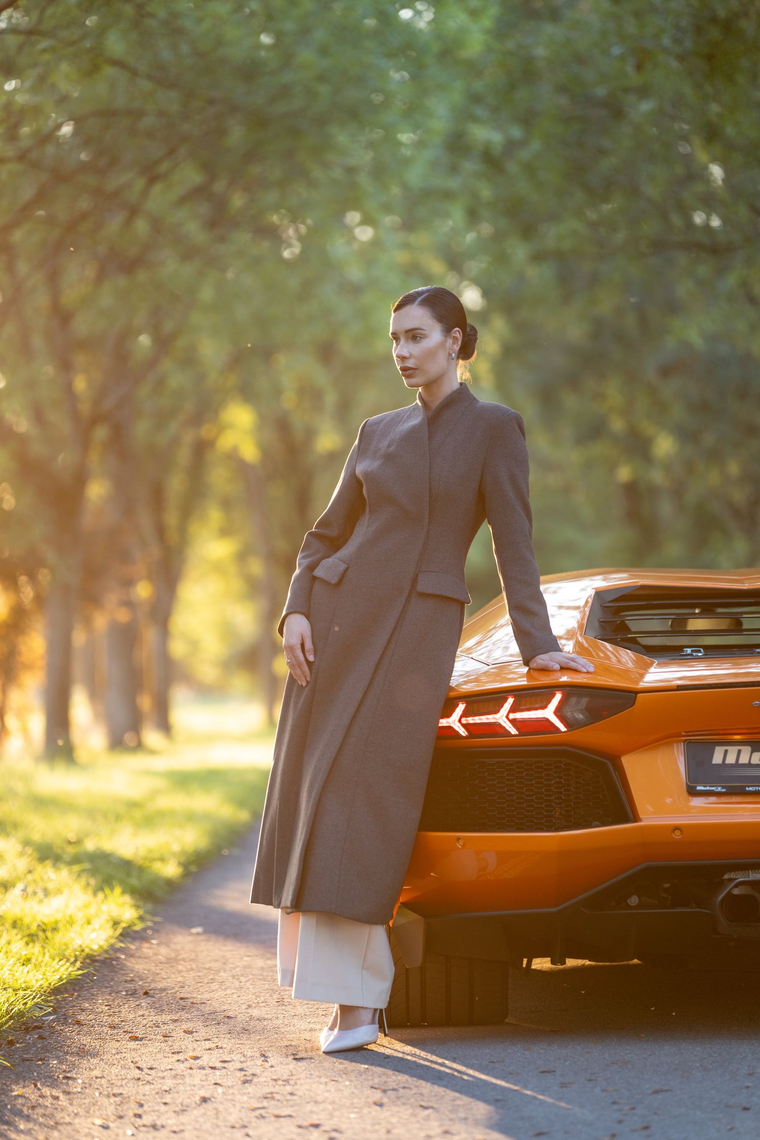 A stylish woman in a long gray coat stands beside an orange Lamborghini Aventador on a tree-lined road.