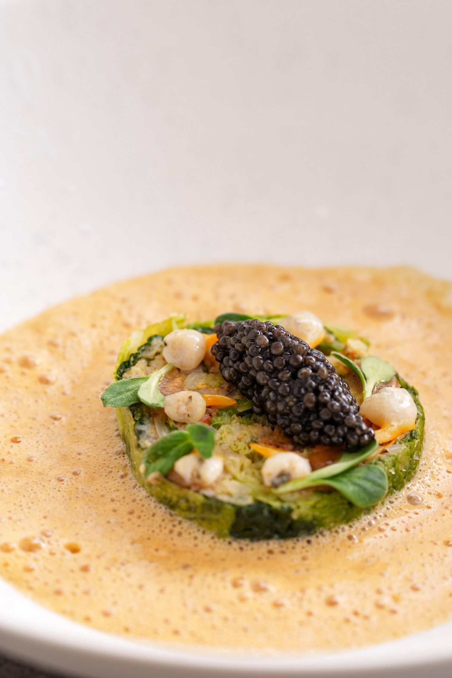 An elegant fine dining dish featuring caviar, microgreens, and pearl garnishes in a frothy golden bisque.
