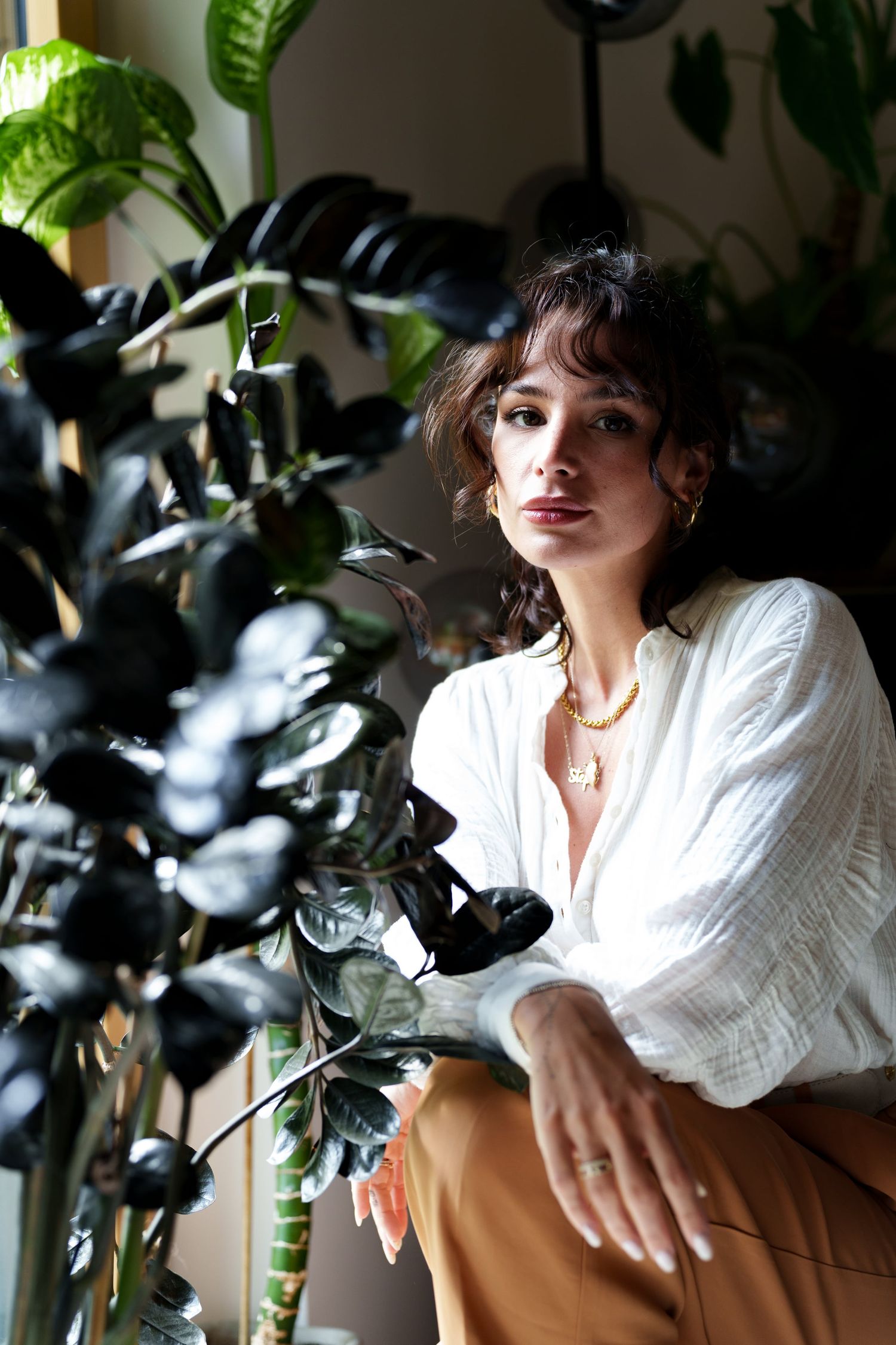 Brunette woman in white blouse sitting among dark tropical plants in indoor setting.