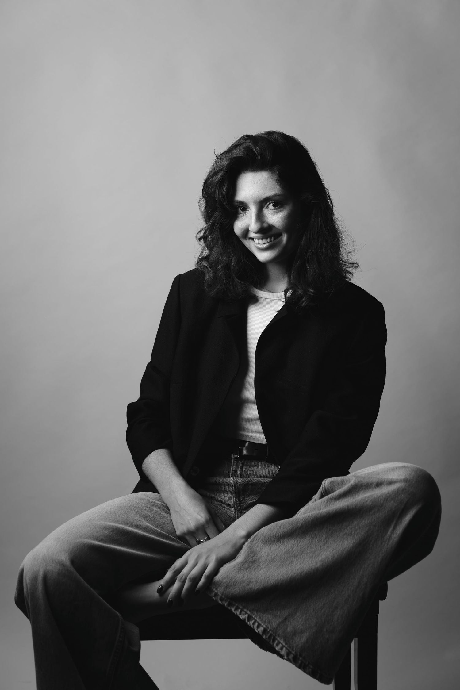Smiling brunette woman in black jacket and jeans sitting on stool in studio.