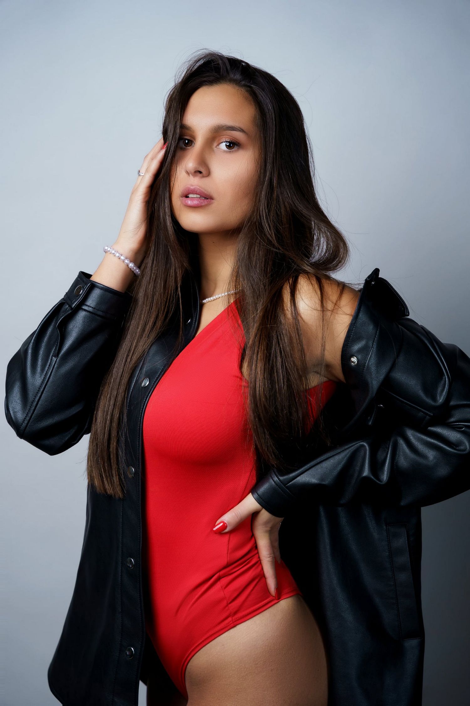 Young brunette woman in a red bodysuit and black leather jacket posing against a gray background.