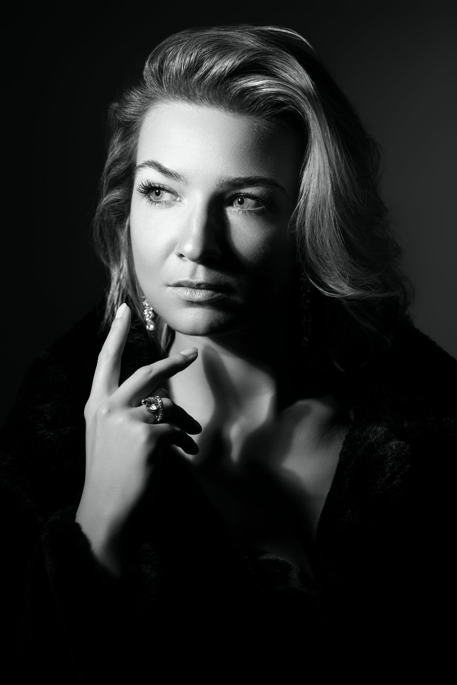 Elegant black and white portrait of blonde woman with jewelry touching her chin in dramatic lighting.