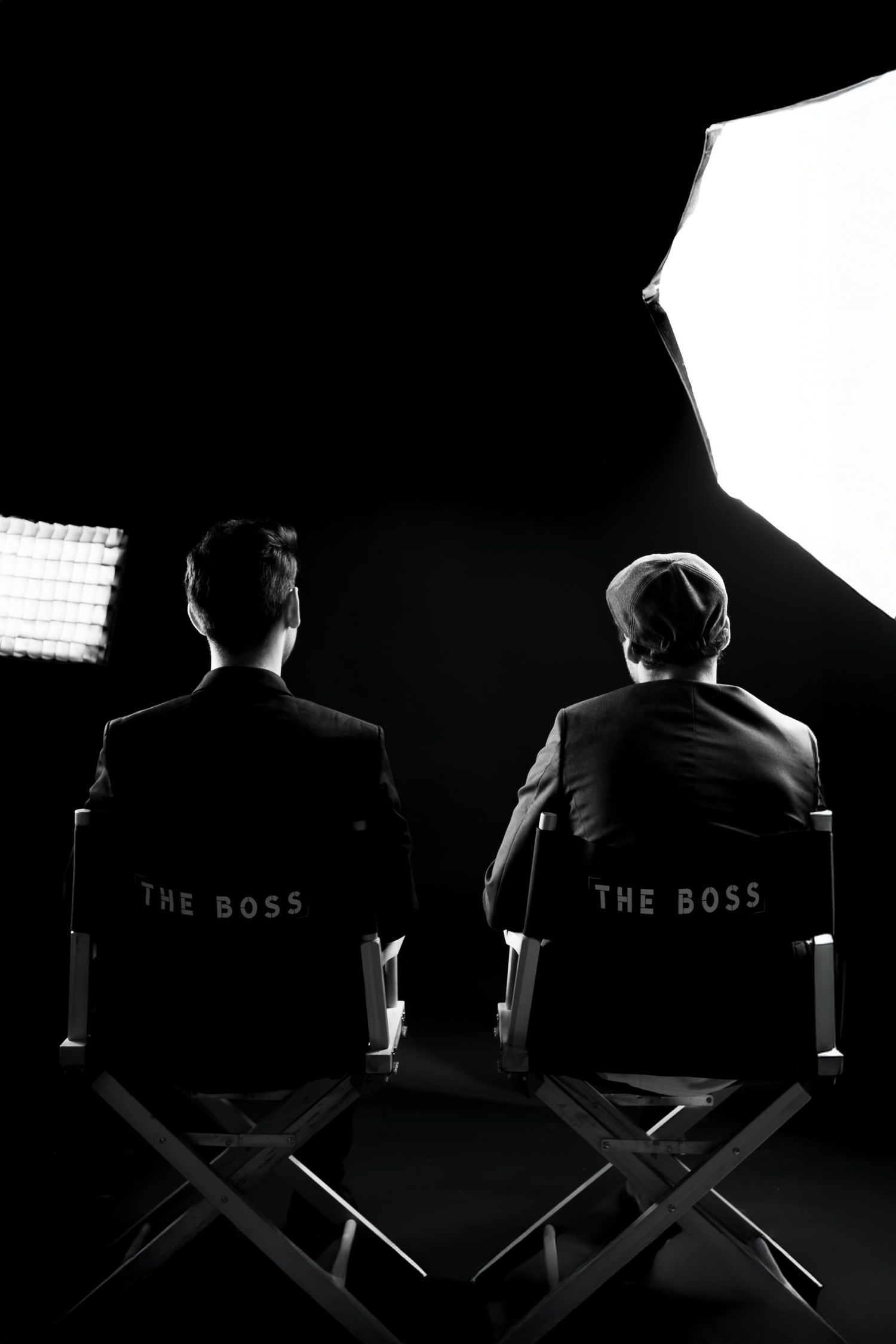 Two men in suits sit in director chairs labeled The Boss in a dramatic black and white studio setting.
