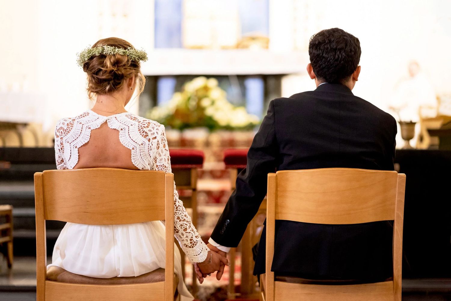 Bride and groom holding hands from behind while seated at church altar during wedding ceremony.