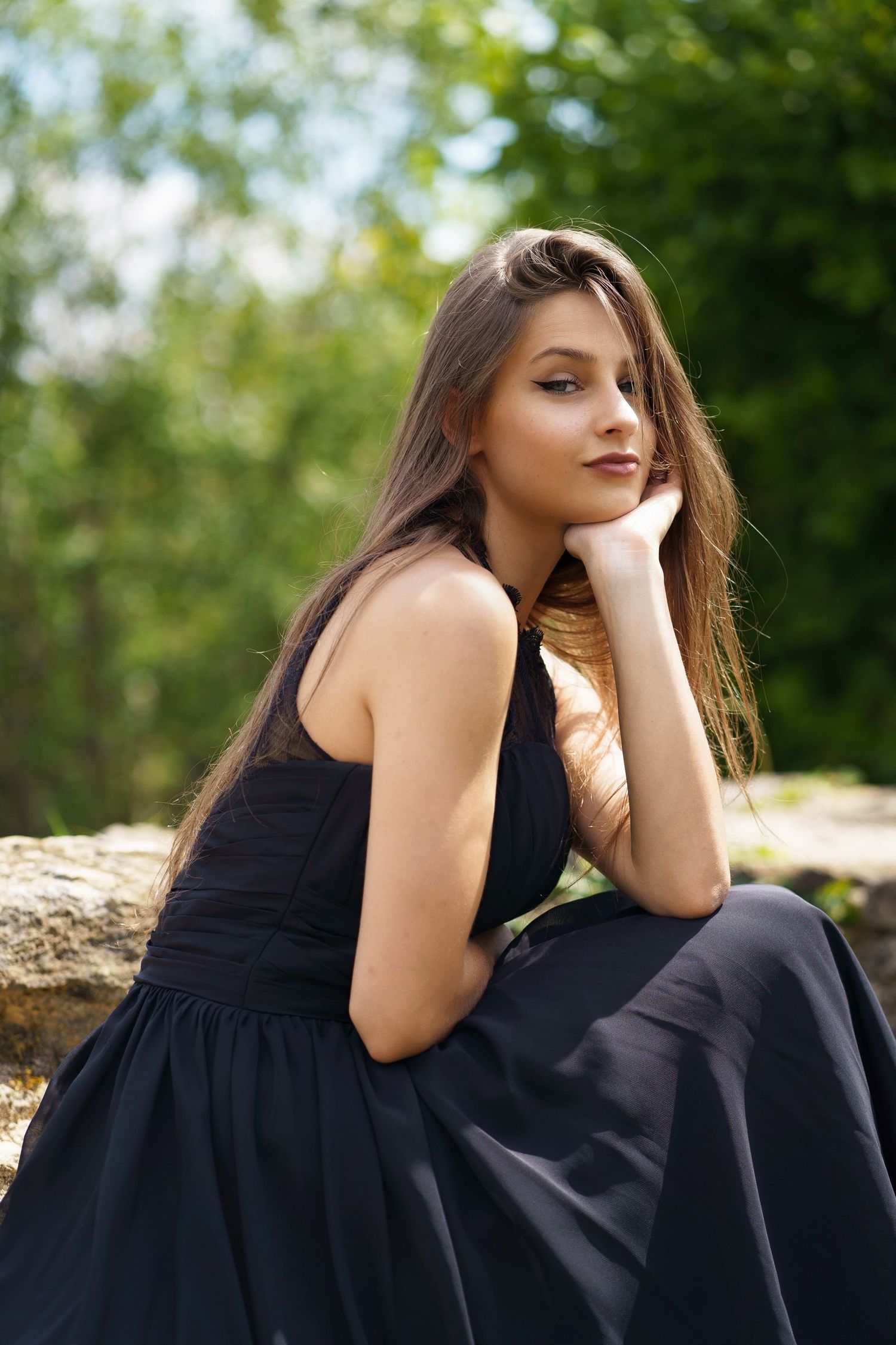 Young woman in black dress sitting on rocks in a lush green outdoor setting.
