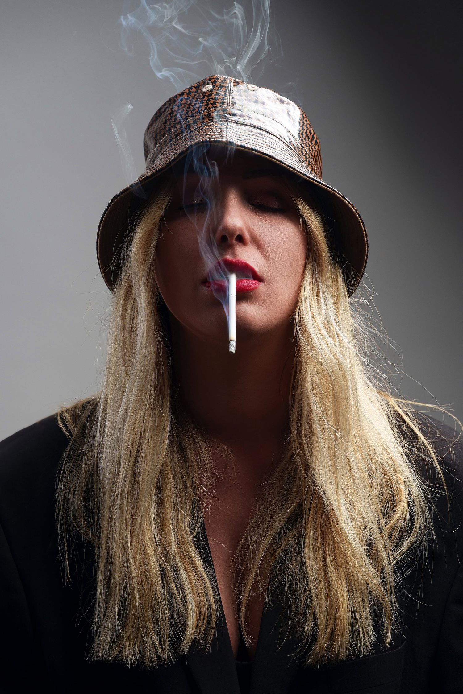 Blonde woman wearing bucket hat smoking cigarette with red lips in studio portrait.