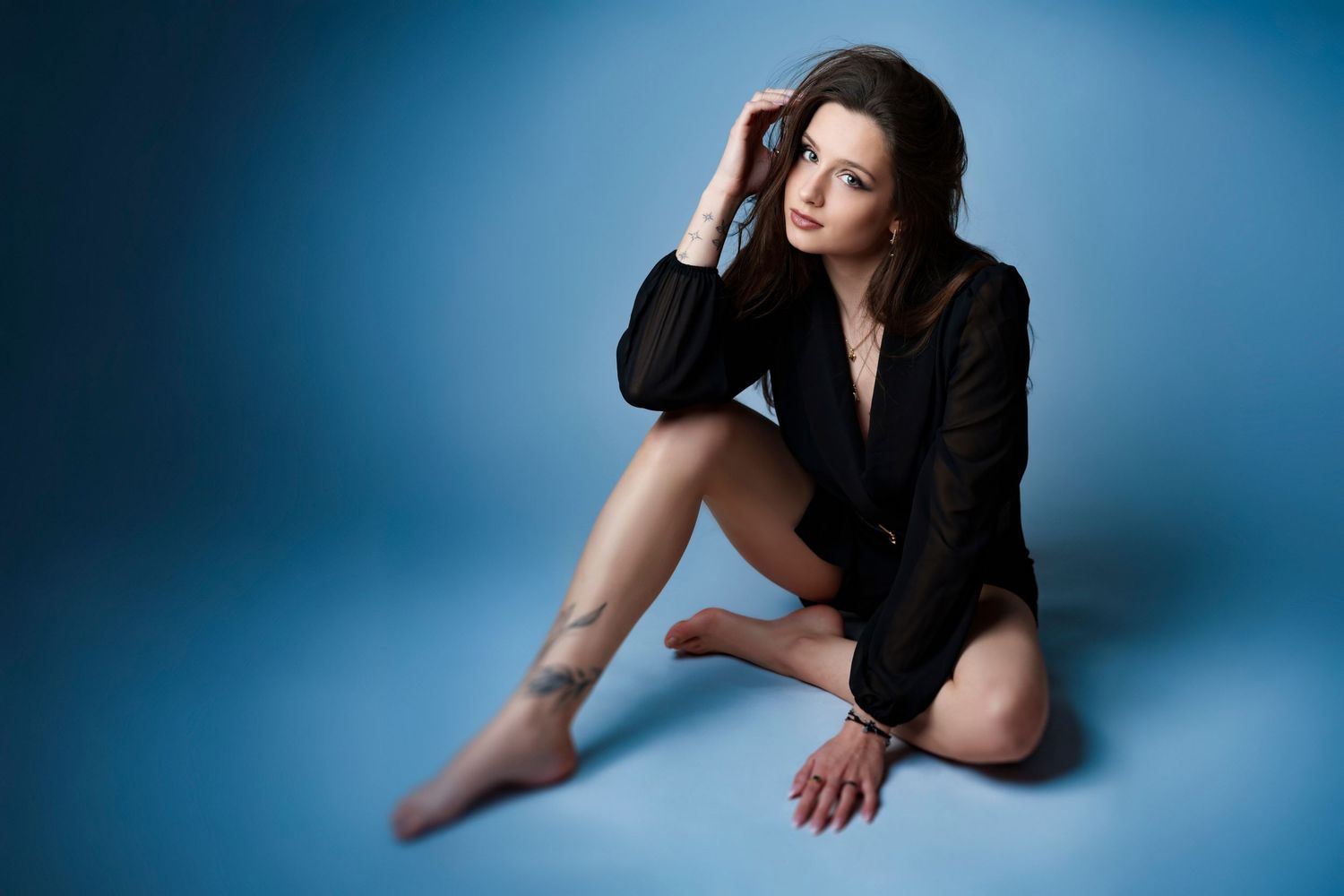 Elegant brunette woman in black blazer sitting gracefully on floor against blue studio background.