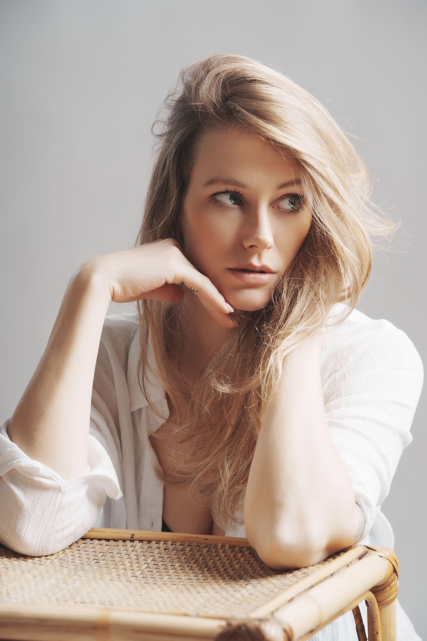 Blonde woman in white shirt resting chin on hand while seated on wicker chair.