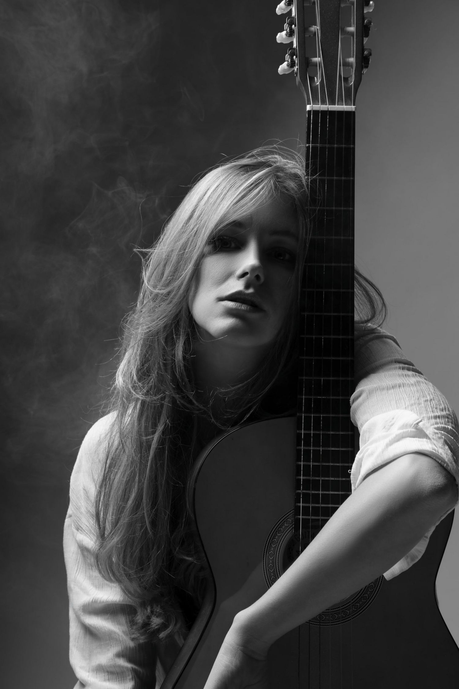 Black and white portrait of a blonde woman leaning against an acoustic guitar in smoky atmosphere.