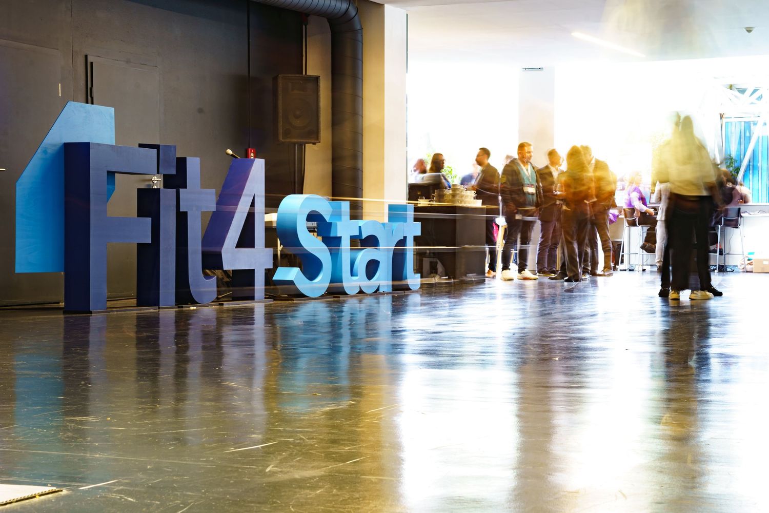 Attendees mingling at Fit4Start startup event in modern convention hall with large blue logo display.