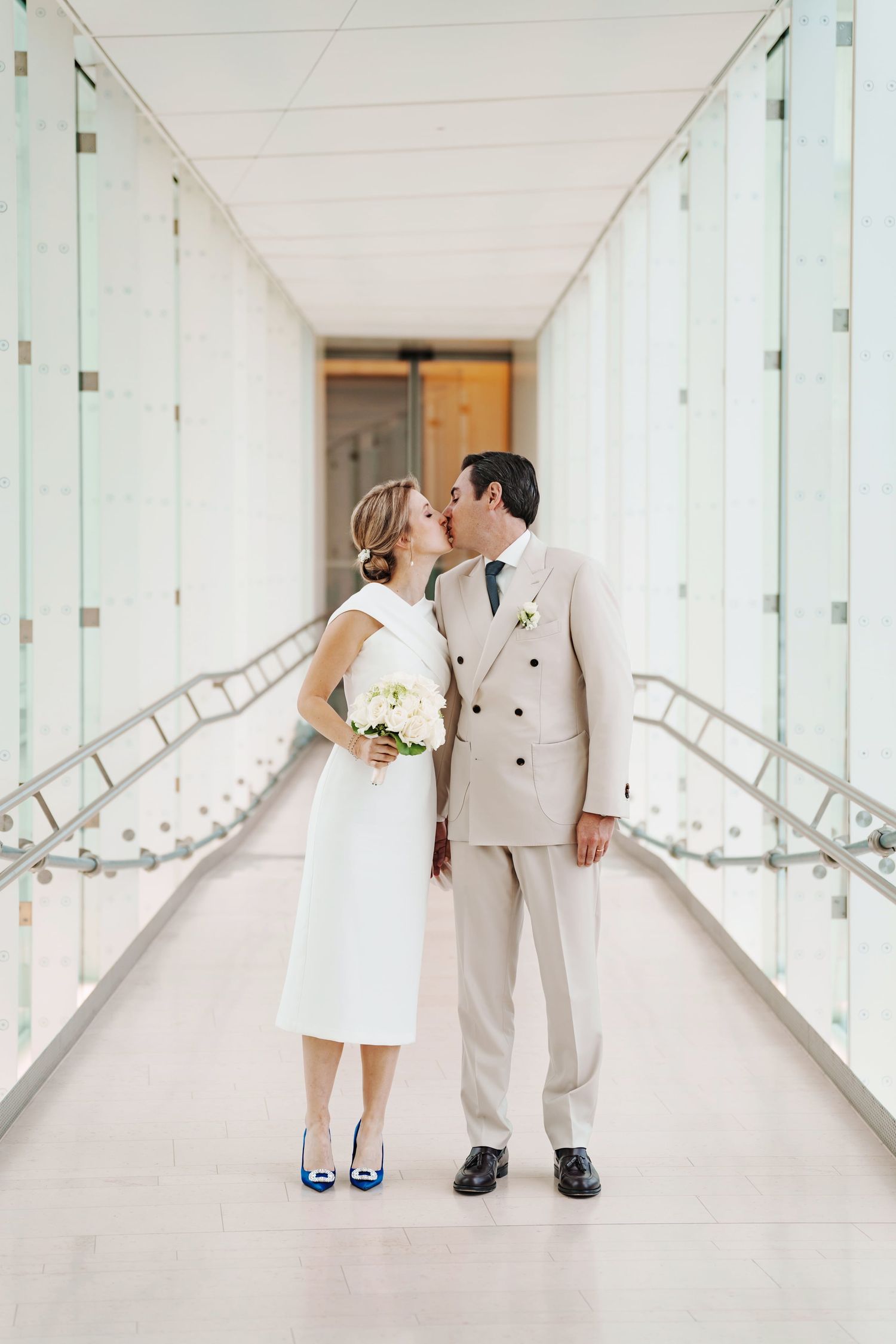 Bride and groom sharing a kiss in a modern white hallway, bride holding white bouquet wearing blue heels.