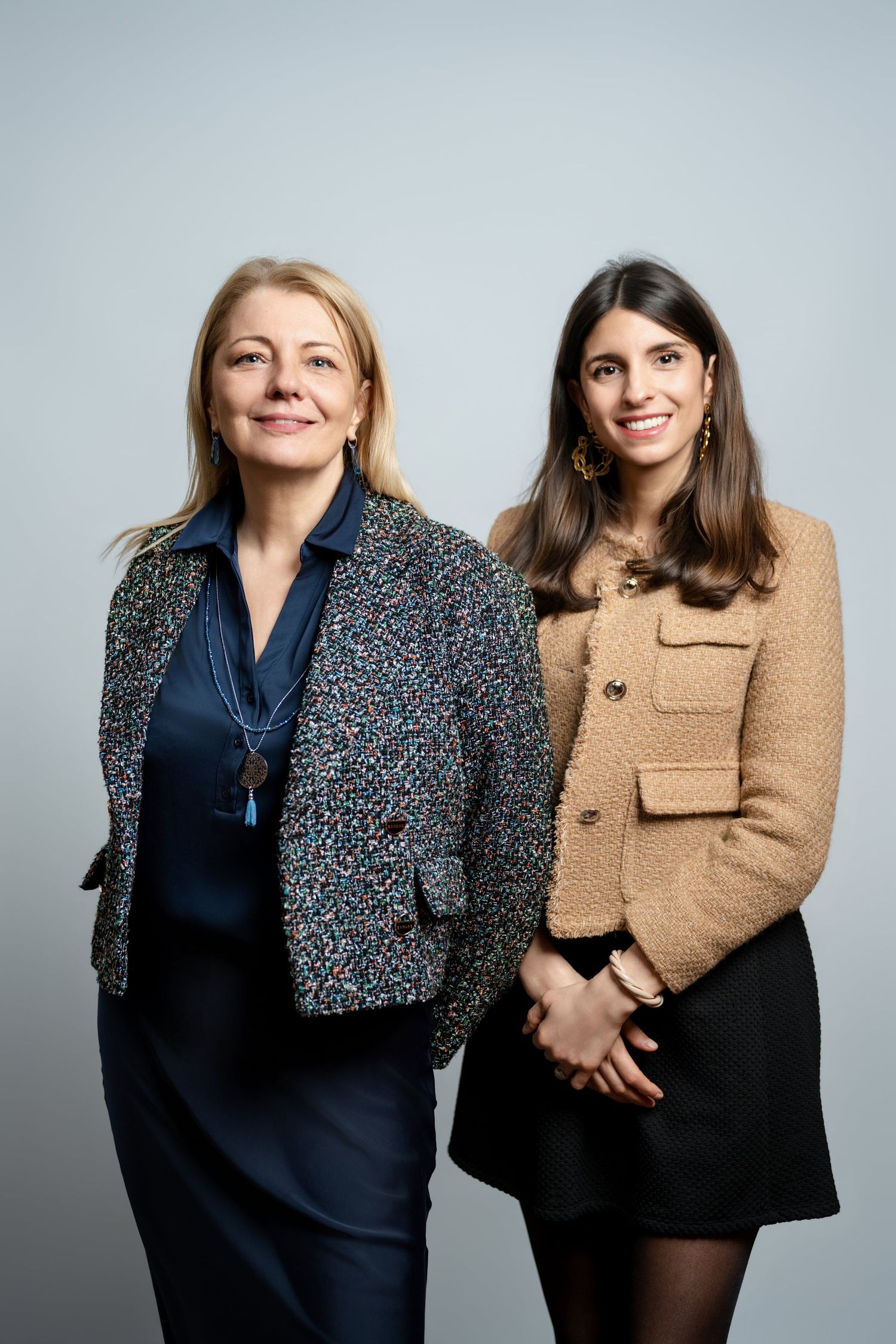 Two professional women smiling in elegant blazers posing together against a light gray studio background.
