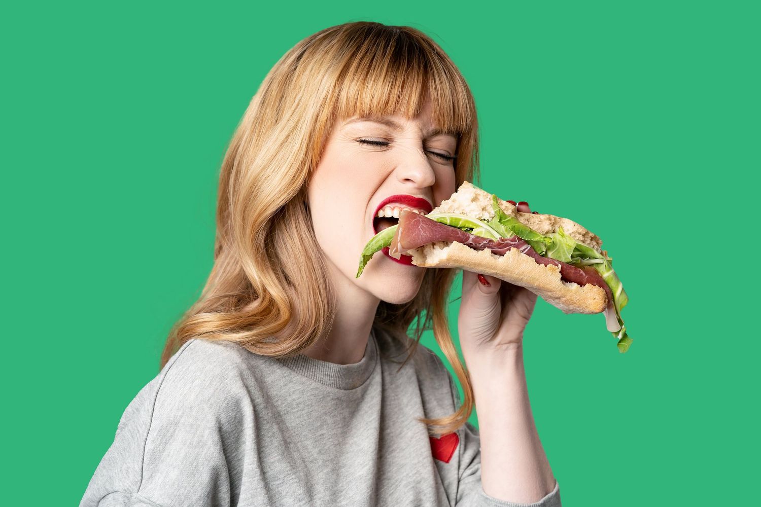 A blonde woman with red lips takes a big bite of a large sandwich against a bright green background.