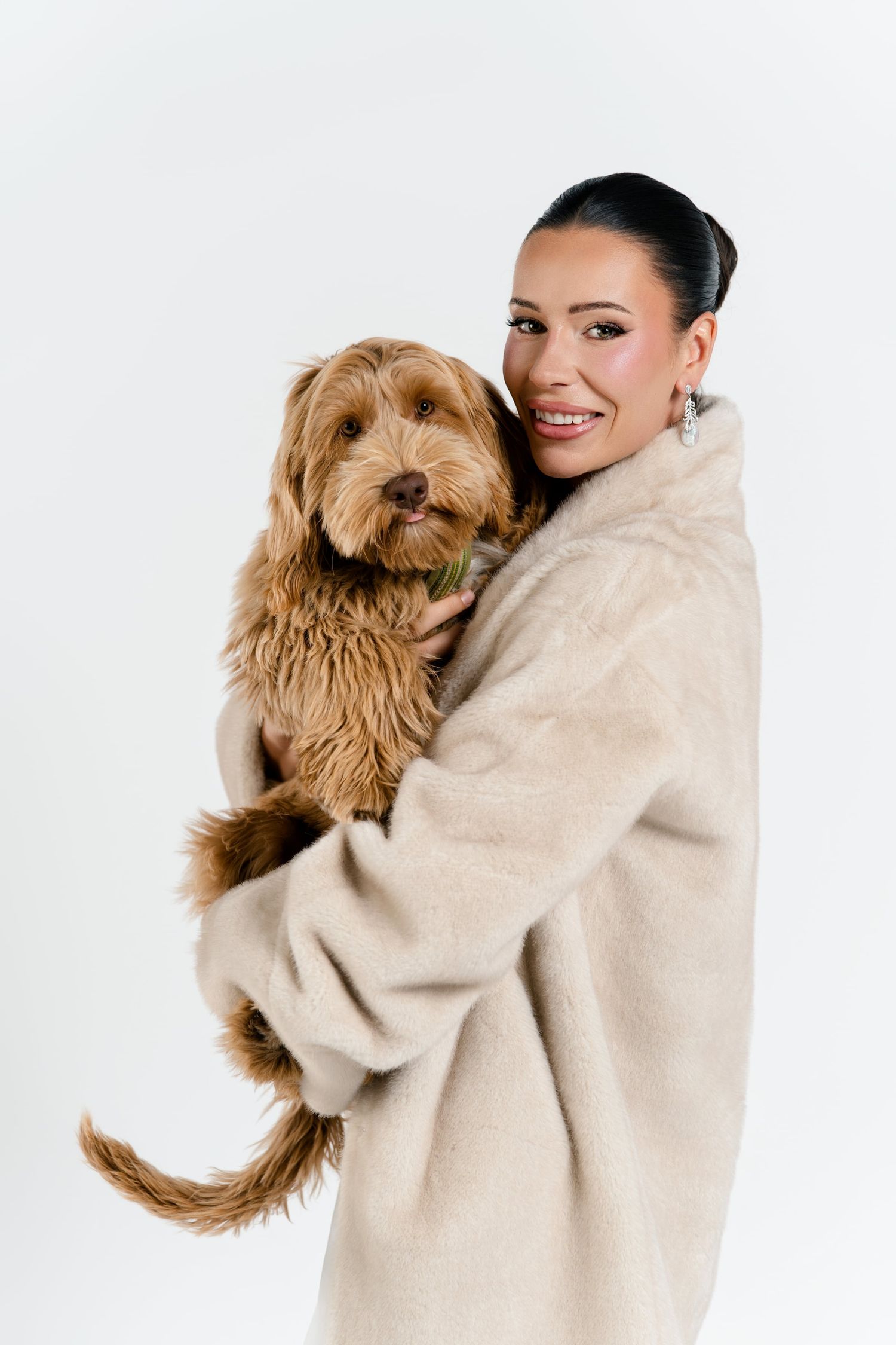 A smiling brunette woman in a beige fur coat holds an adorable fluffy golden Labradoodle dog.