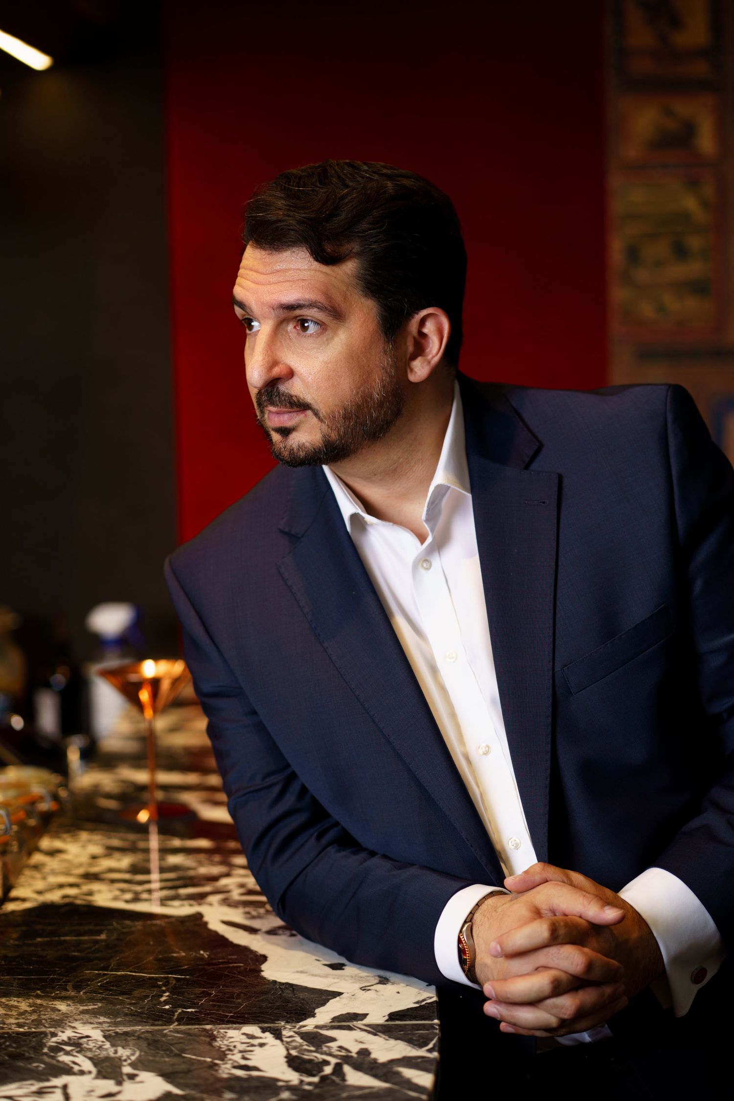 Bearded man in a navy blue suit sitting at a bar counter, looking thoughtfully to the side.