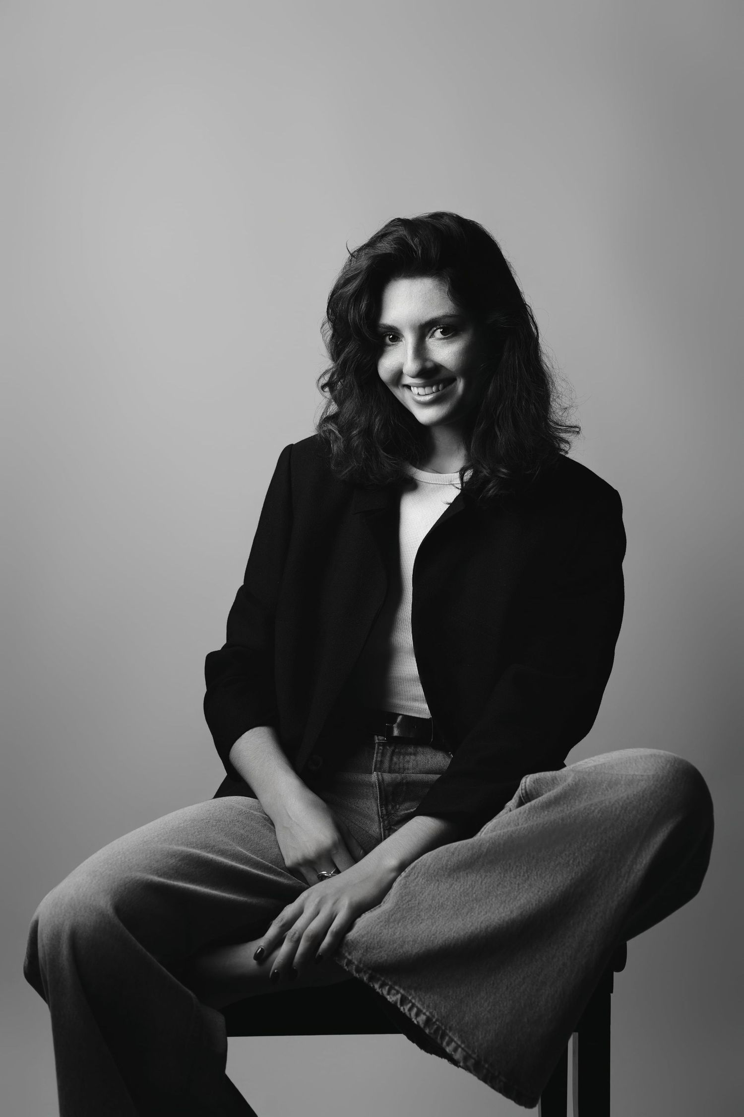 Black and white portrait of smiling brunette woman sitting on stool in casual jacket and jeans.