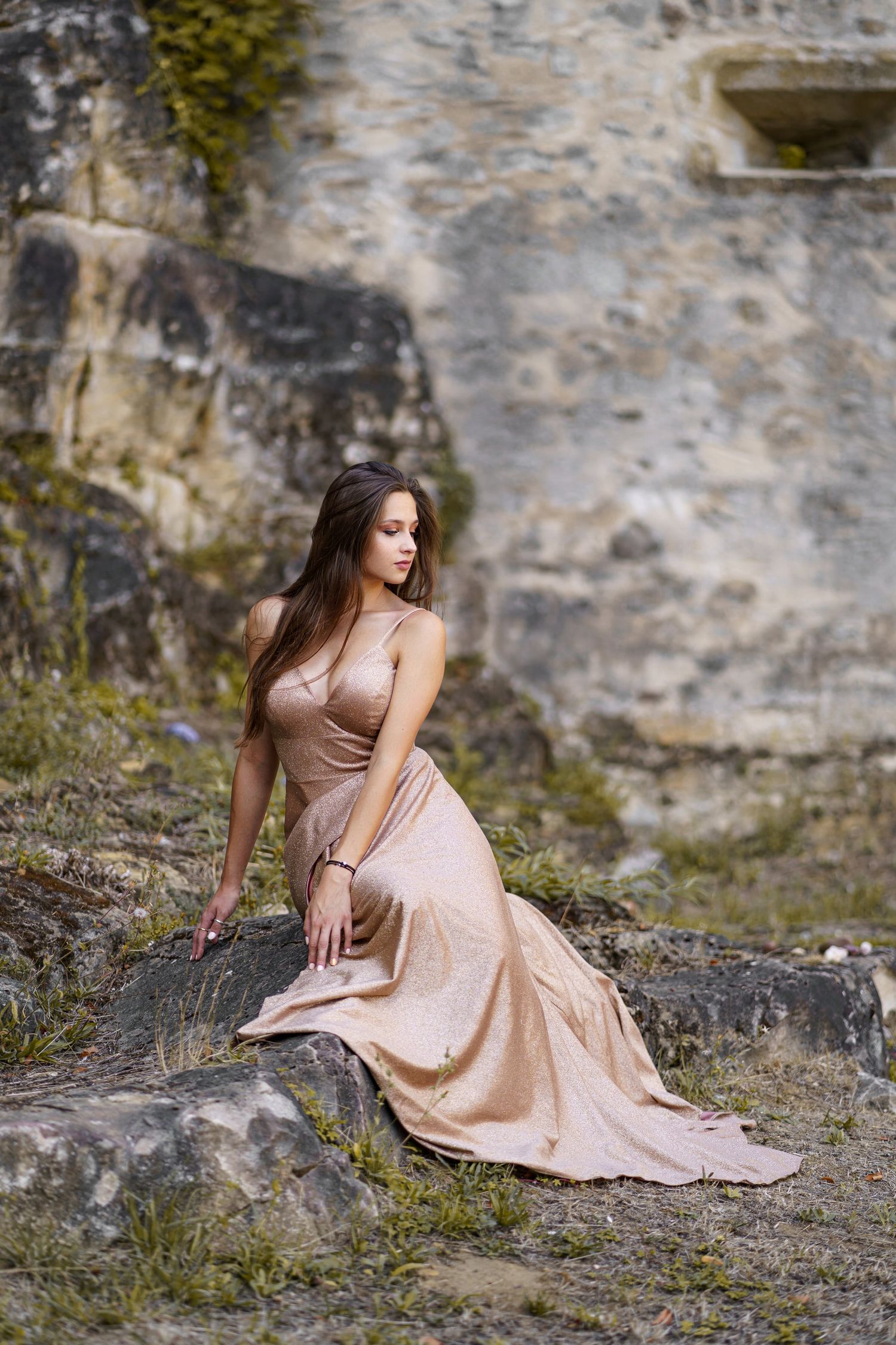 Young woman in elegant gold satin gown sitting on rocks near ancient stone wall outdoors.