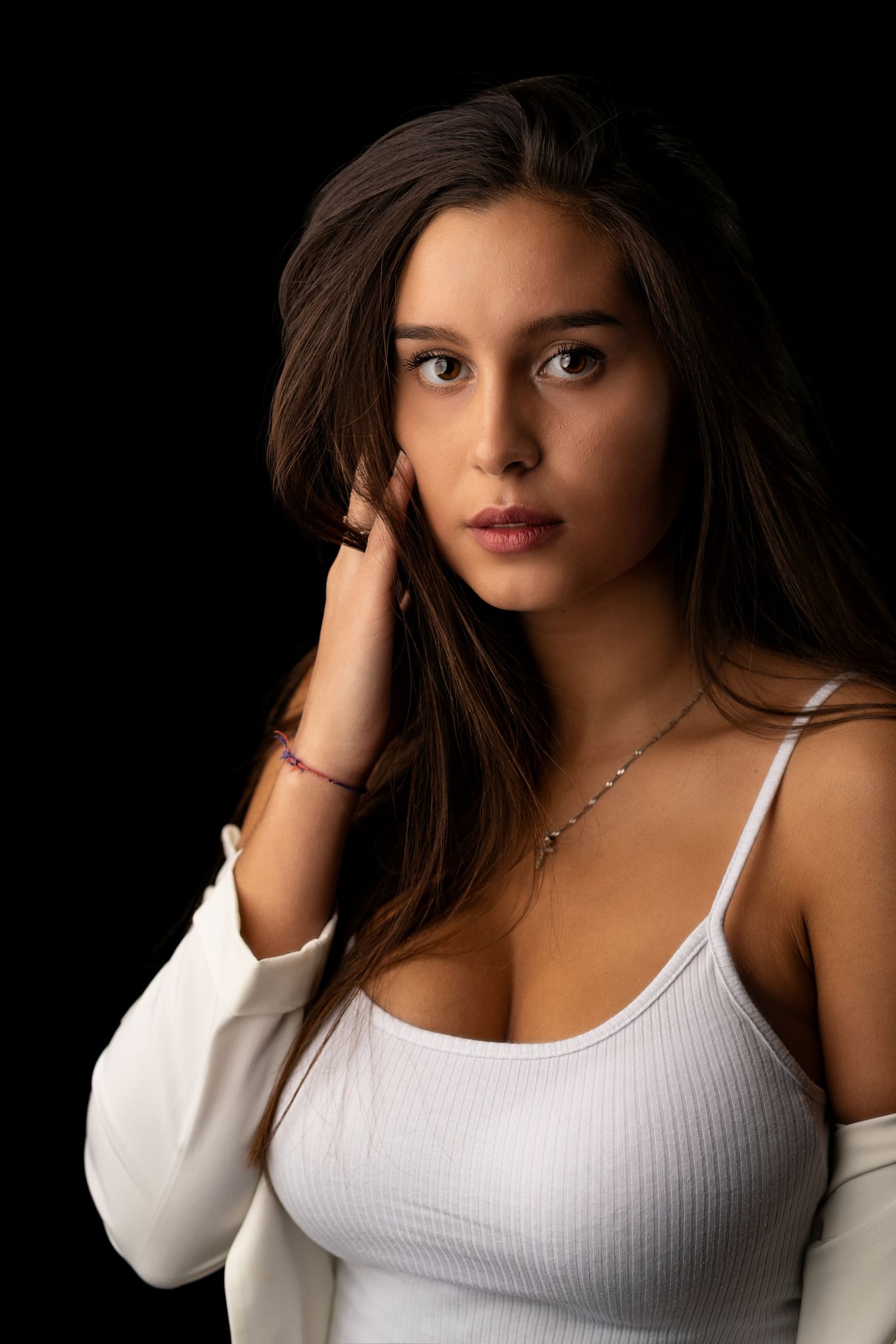 Young brunette woman in white tank top and jacket posing against a dark studio background.