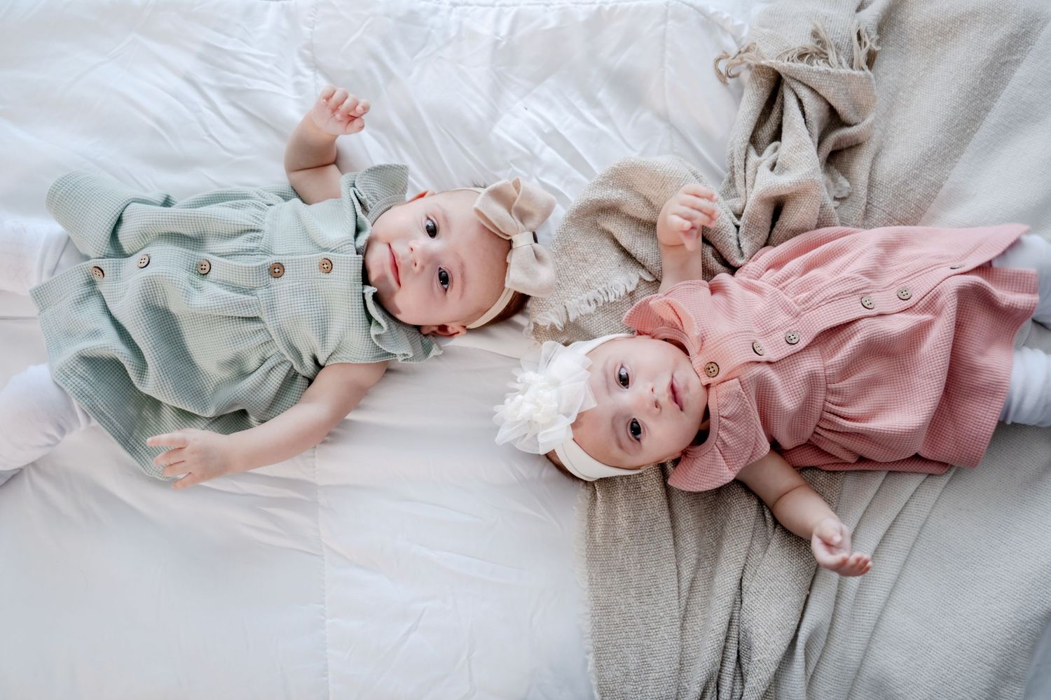 Two babies in pastel dresses lying on white bedding smiling.