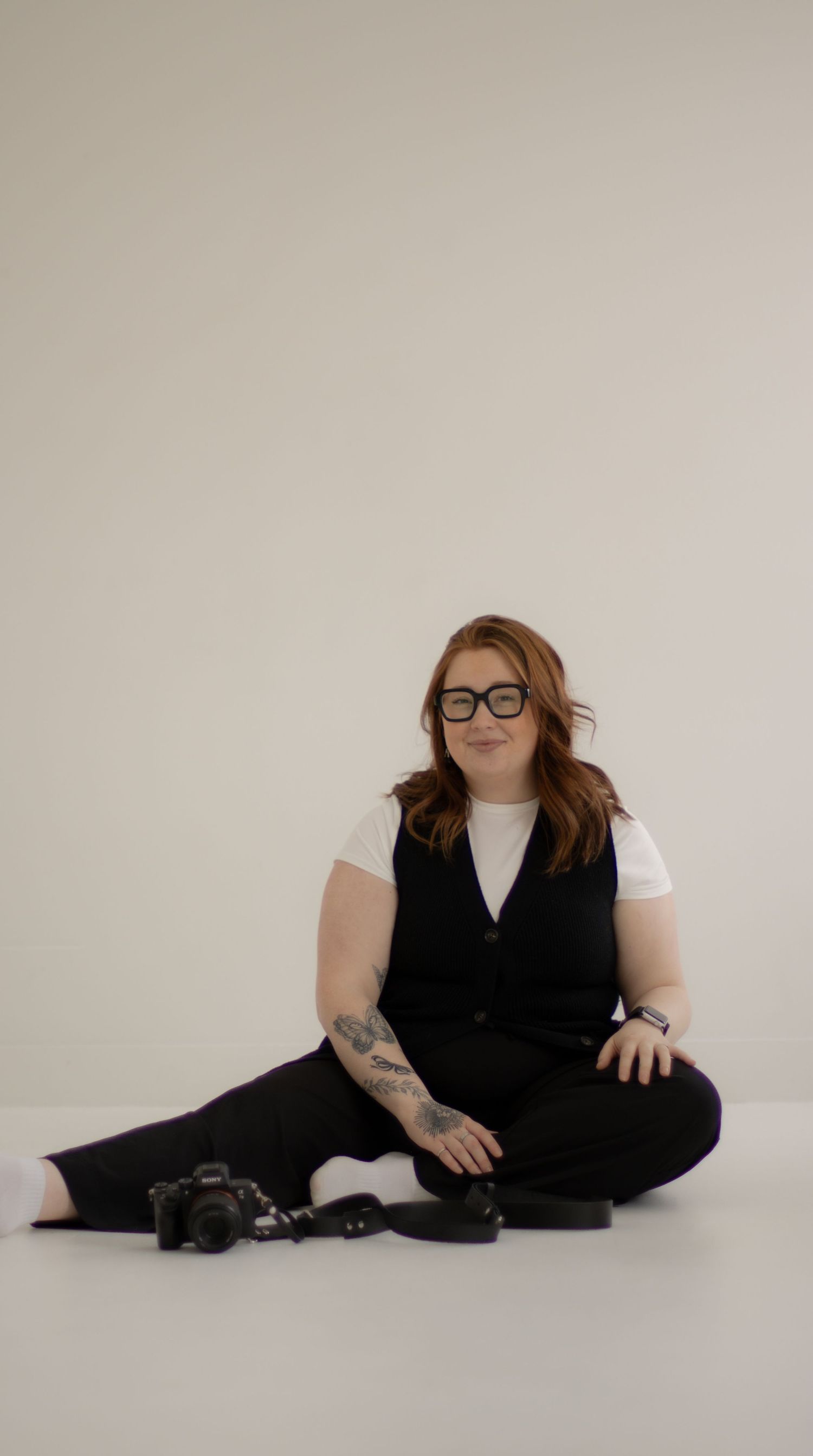 A creative professional in glasses sits on the floor wearing black vest and pants with visible tattoos.