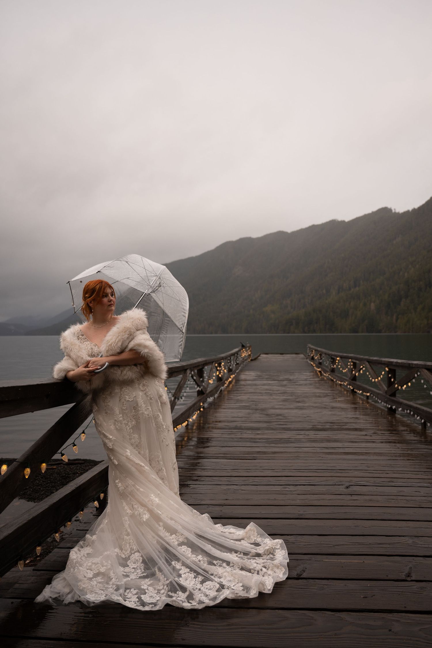 A bride leans against the railing of a dock and holds an umbrella against the rain on a moody PNW day.