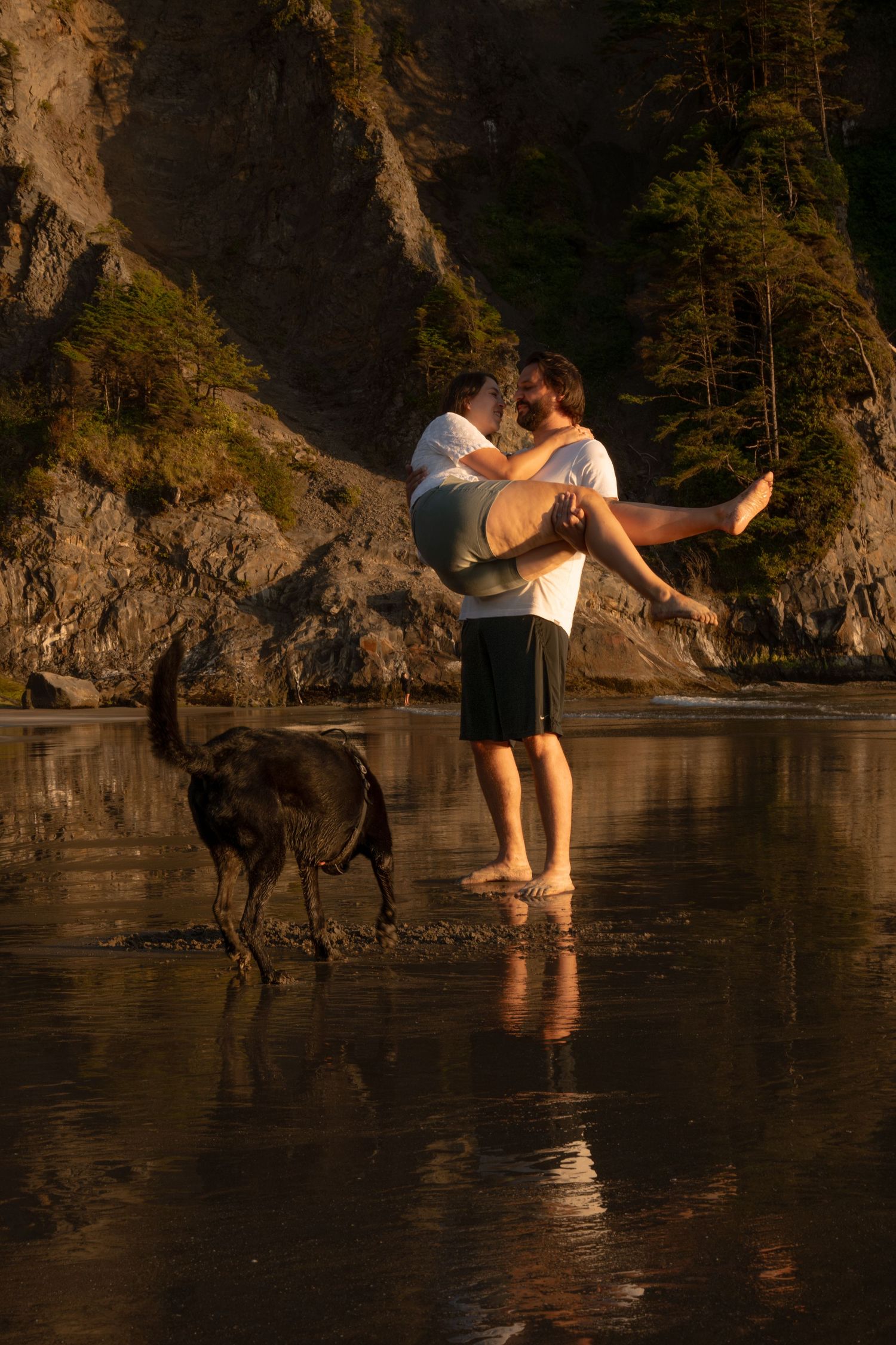 A young man holds his wife on a beach during sunset while their black lab plays near by on the norther Oregon coast.