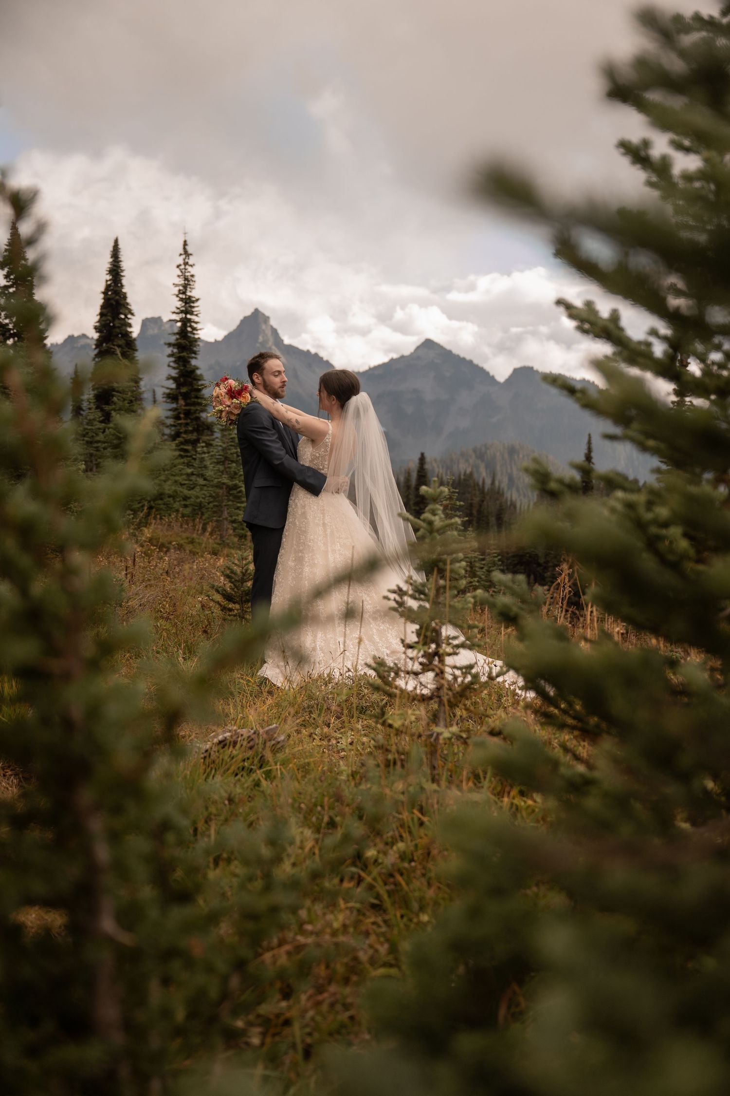 A bridge and groom embrace in an alpine meadow with mountains in the distance.