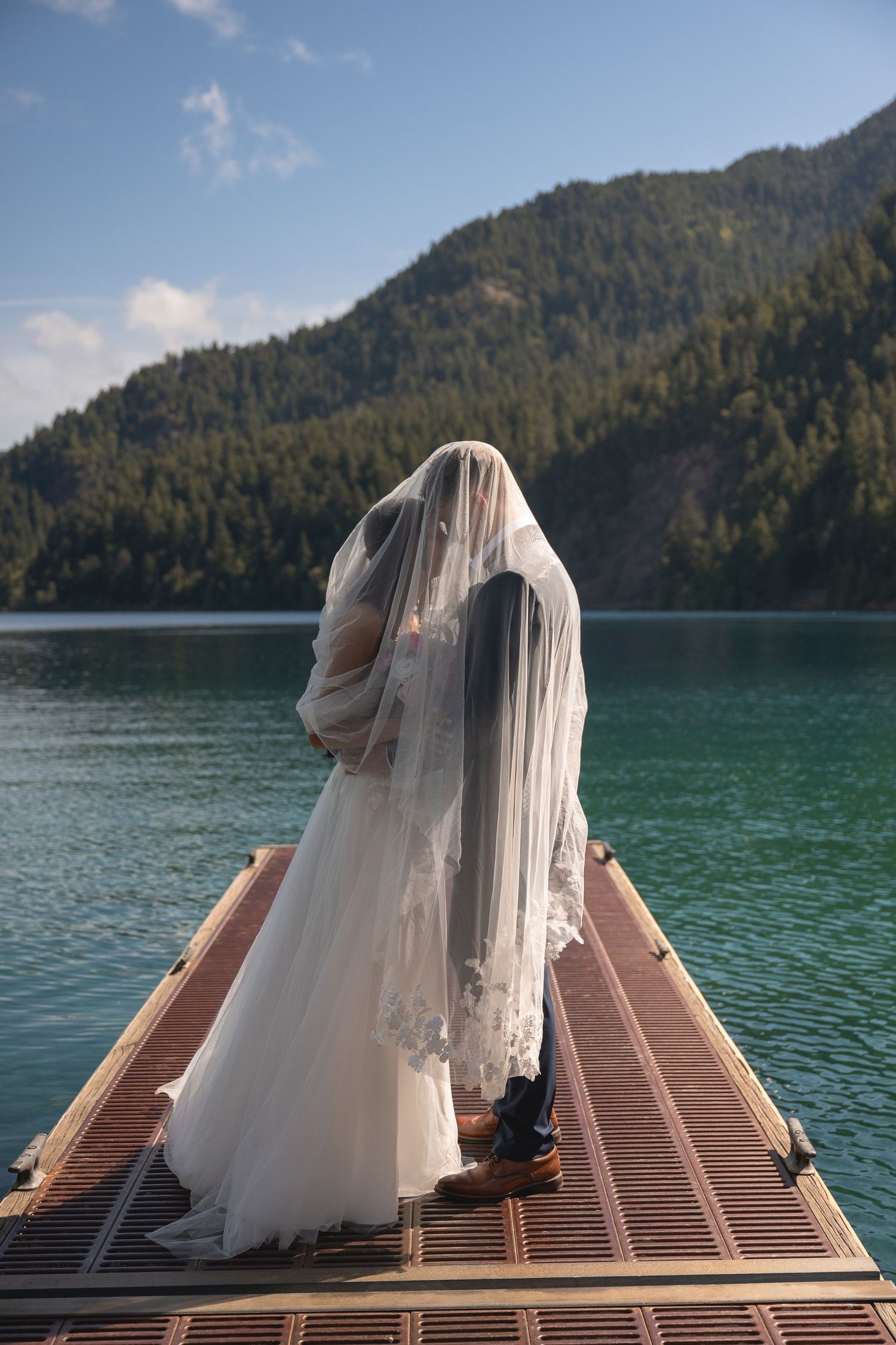 A veil covers an eloping couple standing at the end of a dock on Lake Crescent in Olympic National Park.