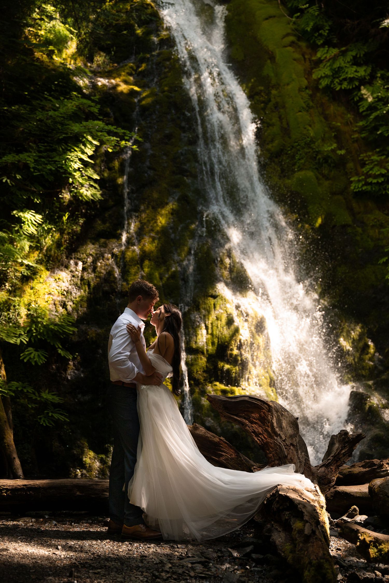 A young couple in wedding attire kisses in front of an Olympic National Park waterfall (Marymere Falls).