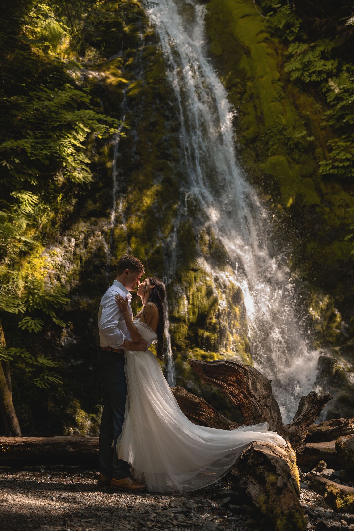 A young couple in wedding attire kisses in front of an Olympic National Park waterfall (Marymere Falls).