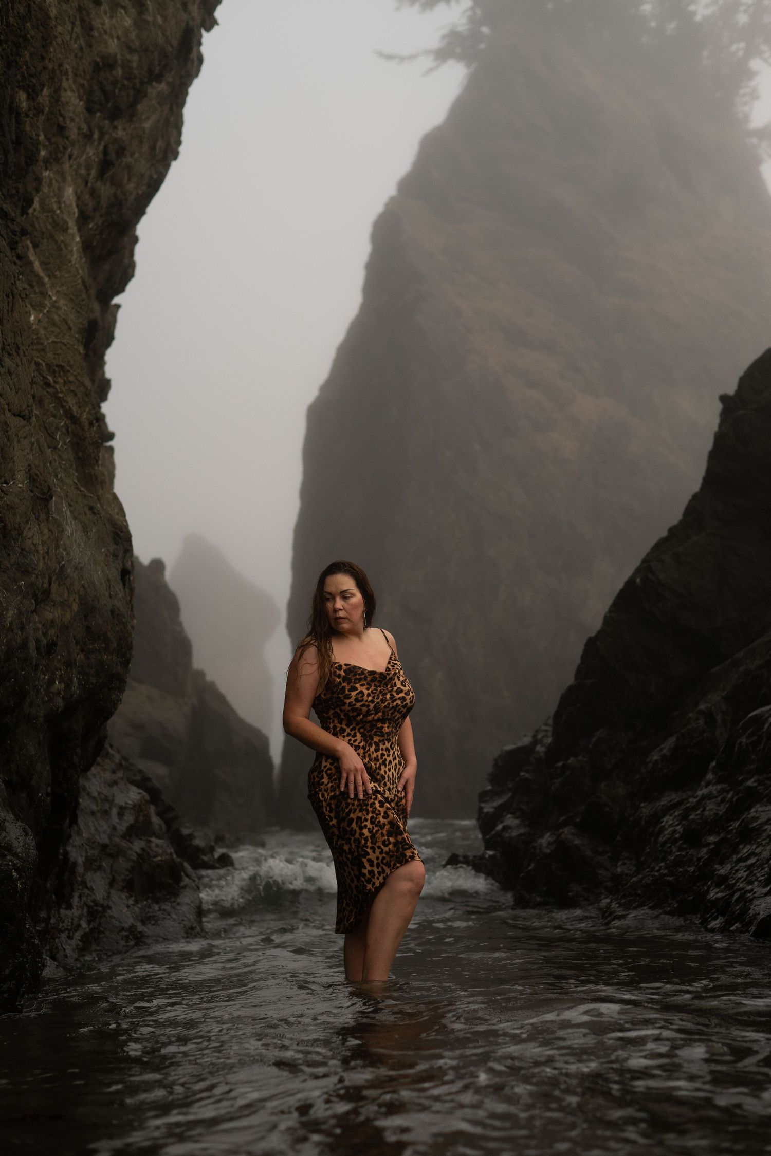 A young woman poses in shallow water on a foggy and moody day at Secret Beach on the Southern Oregon coast.