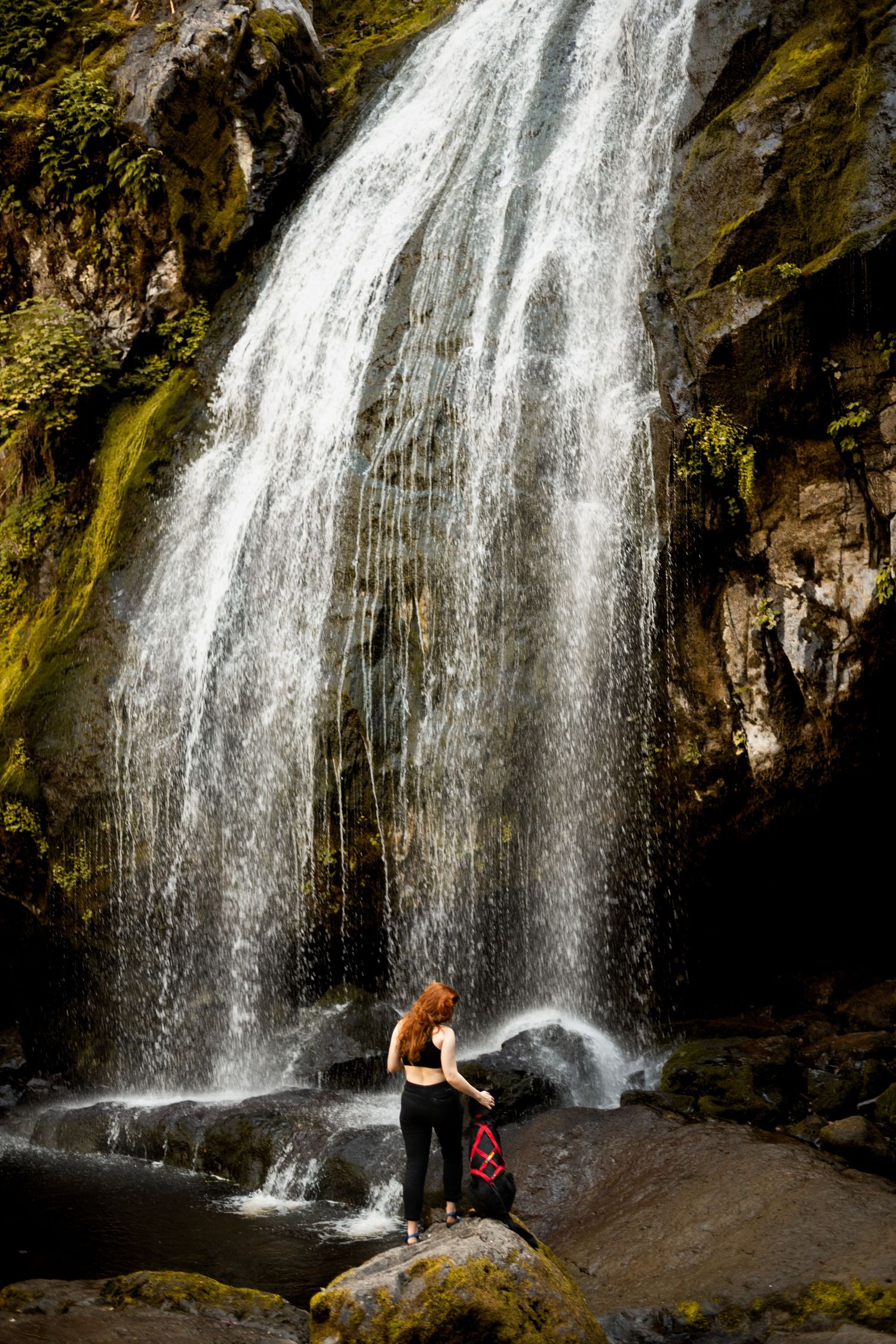 A young woman poses with her black German shepherd in front of a tall waterfall near Mt. Rainier in western Washington.