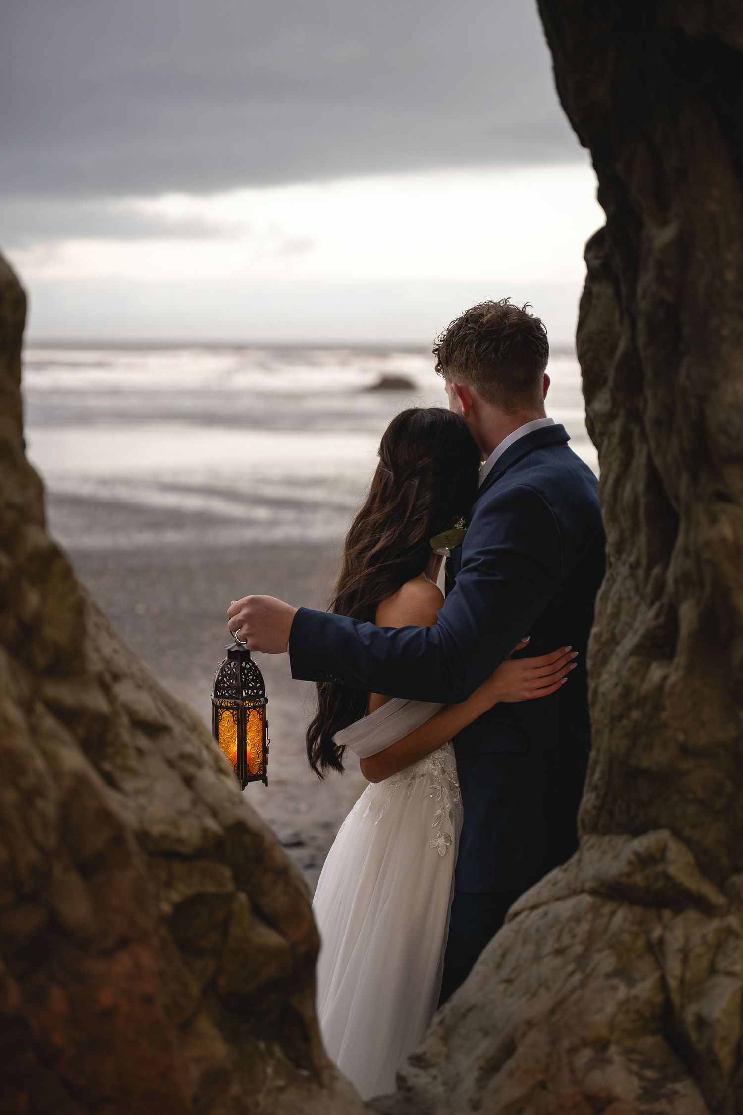 A groom embraces his bride in front of sea stacks on Ruby Beach in Olympic National Park while holding lantern to light the early morning.