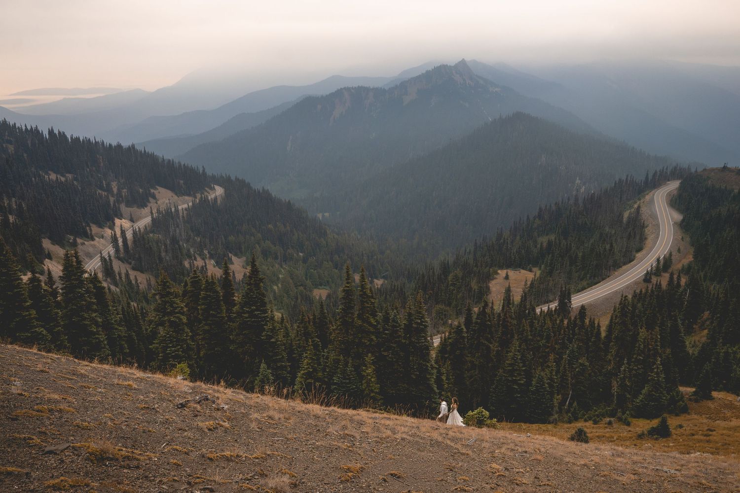 A couple hikes in their wedding attire along a trail near Hurricane Ridge on a hazy summer day in Washington.