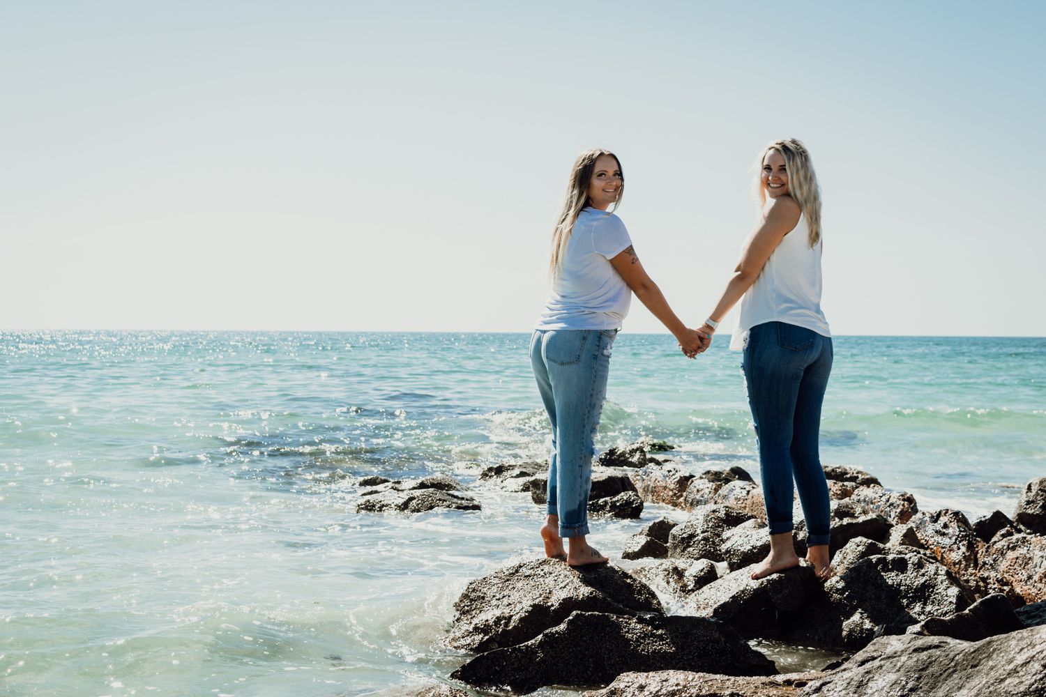 Two best friends hold hands while walking along a rocky beach shoreline with turquoise ocean waters in the background.