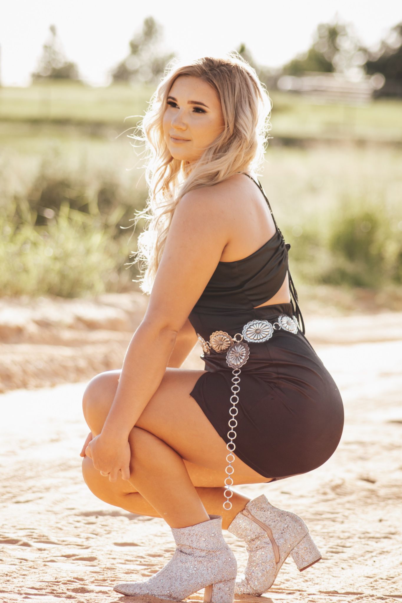 A woman in a black dress and white boots poses on sandy ground in warm sunlight.