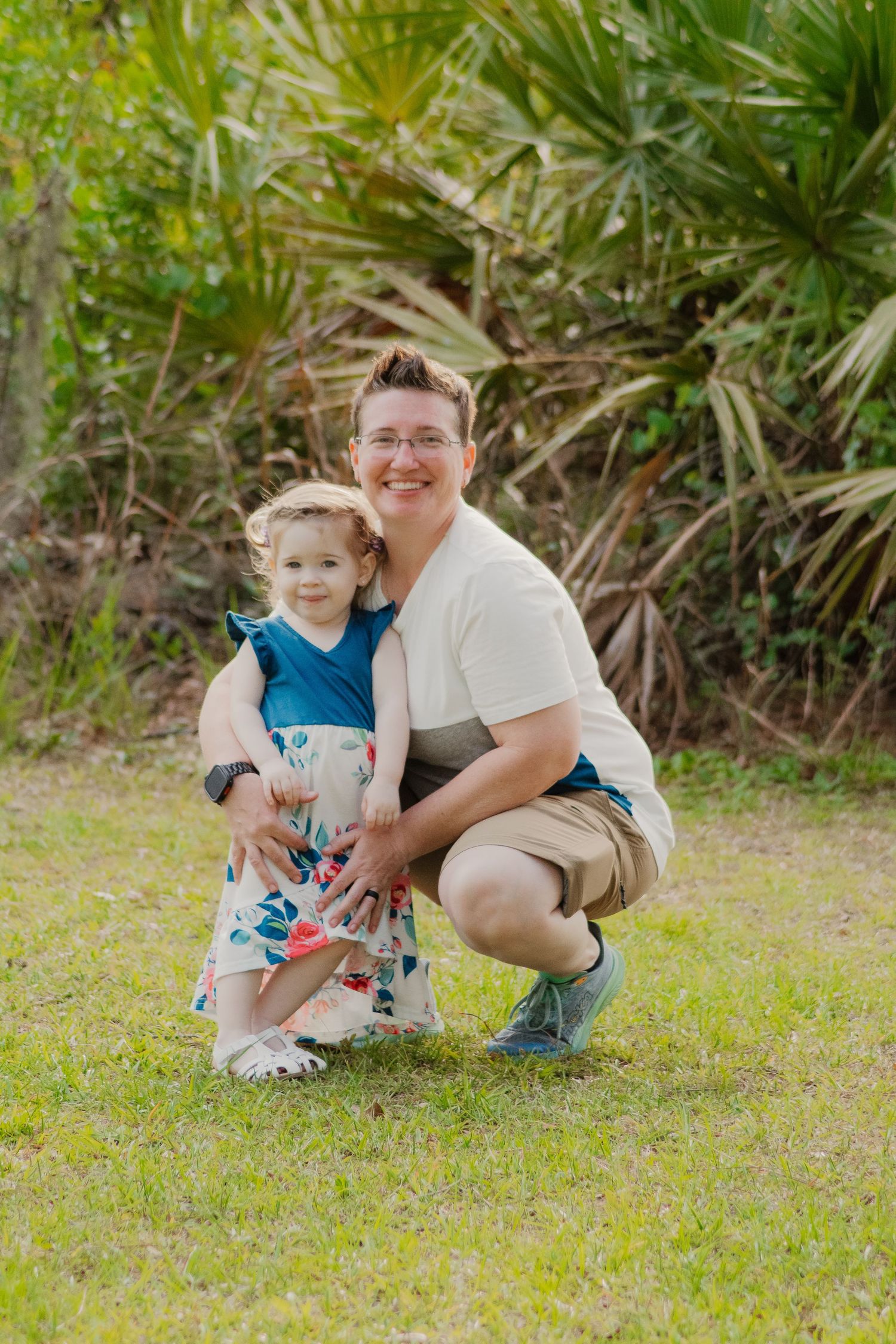 Parent and child share a tender moment together sitting on grass surrounded by tropical plants and greenery.