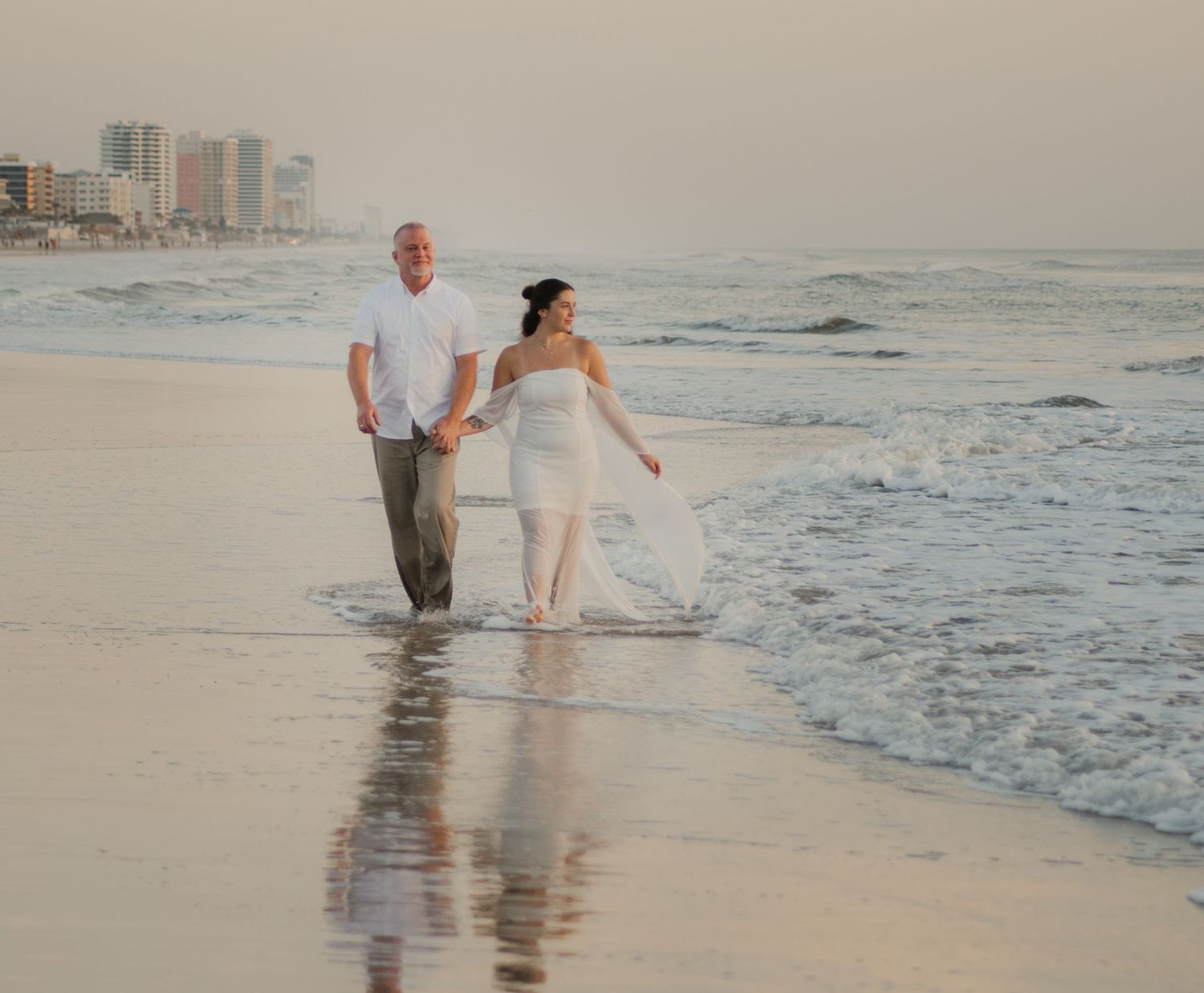 A romantic couple walks on the beach at sunset near a wooden pier.