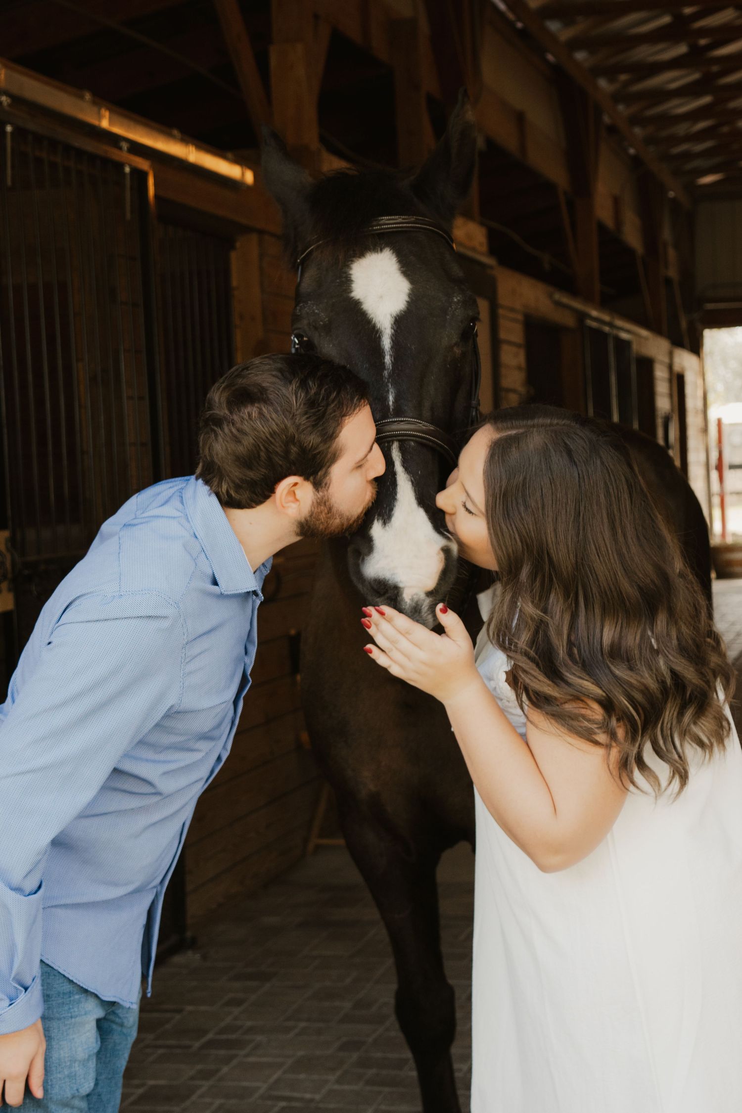 A couple shares a playful kissing moment with a black horse with white markings in a rustic barn stable.
