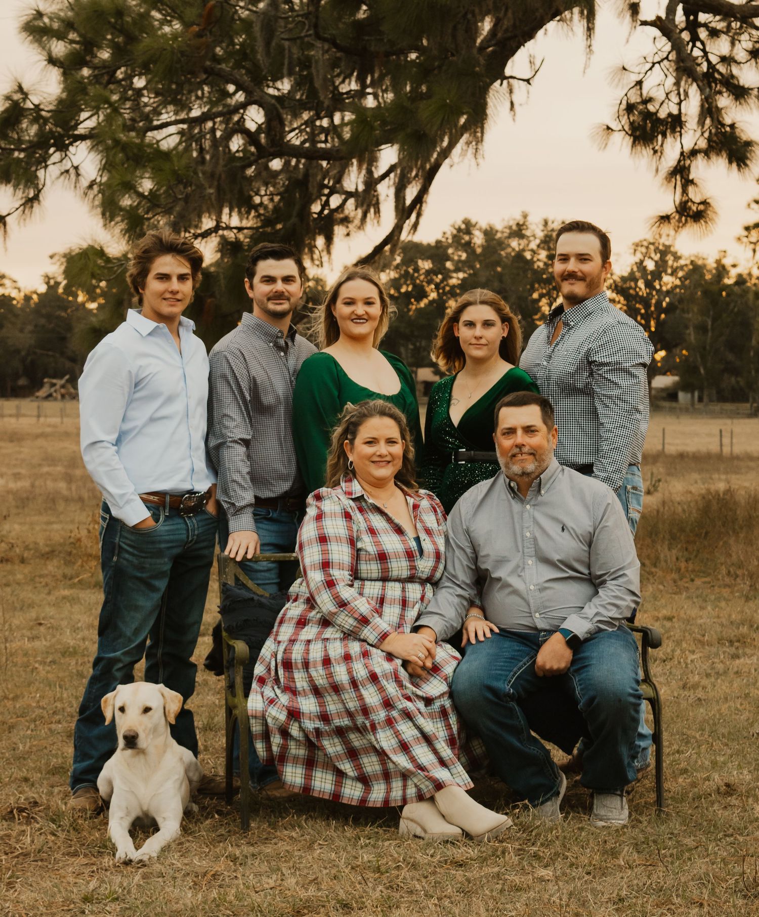 A family in coordinating blue and green outfits poses together on grass with their lab at sunset.