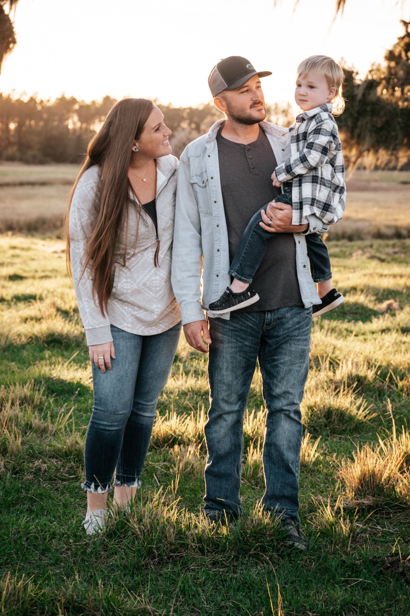 Family walking together in a grassy field at sunset wearing casual clothing and jeans.